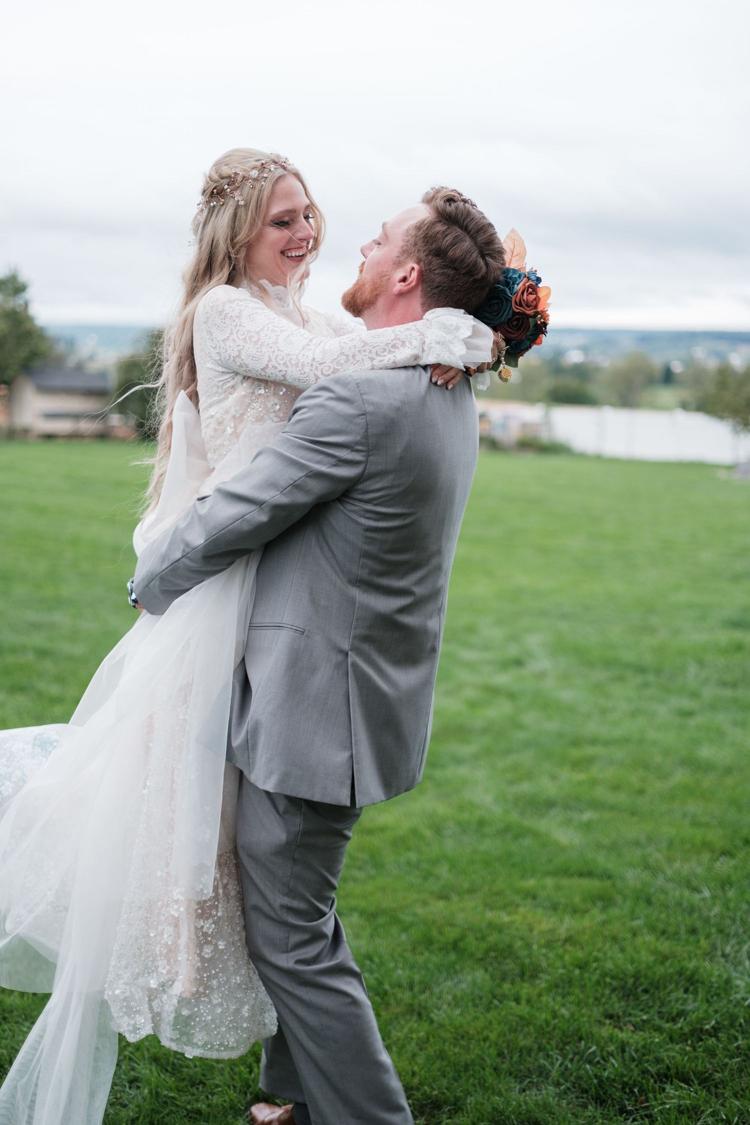 Groom in a gray suit lifts his smiling bride in a lace wedding gown on a green lawn during an outdoor wedding photo session.