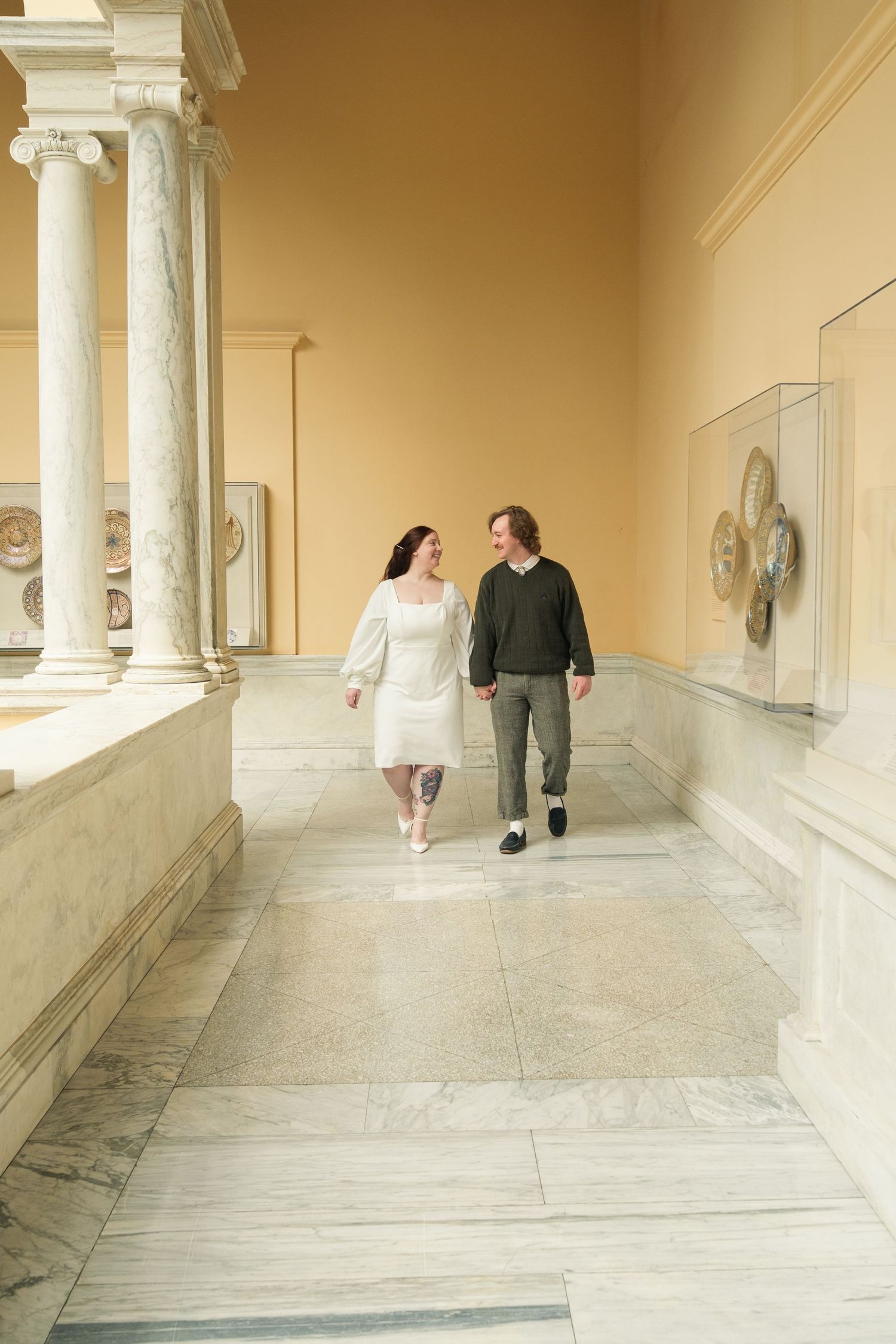 Couple holding hands walking through elegant museum hallway with marble columns and yellow walls.