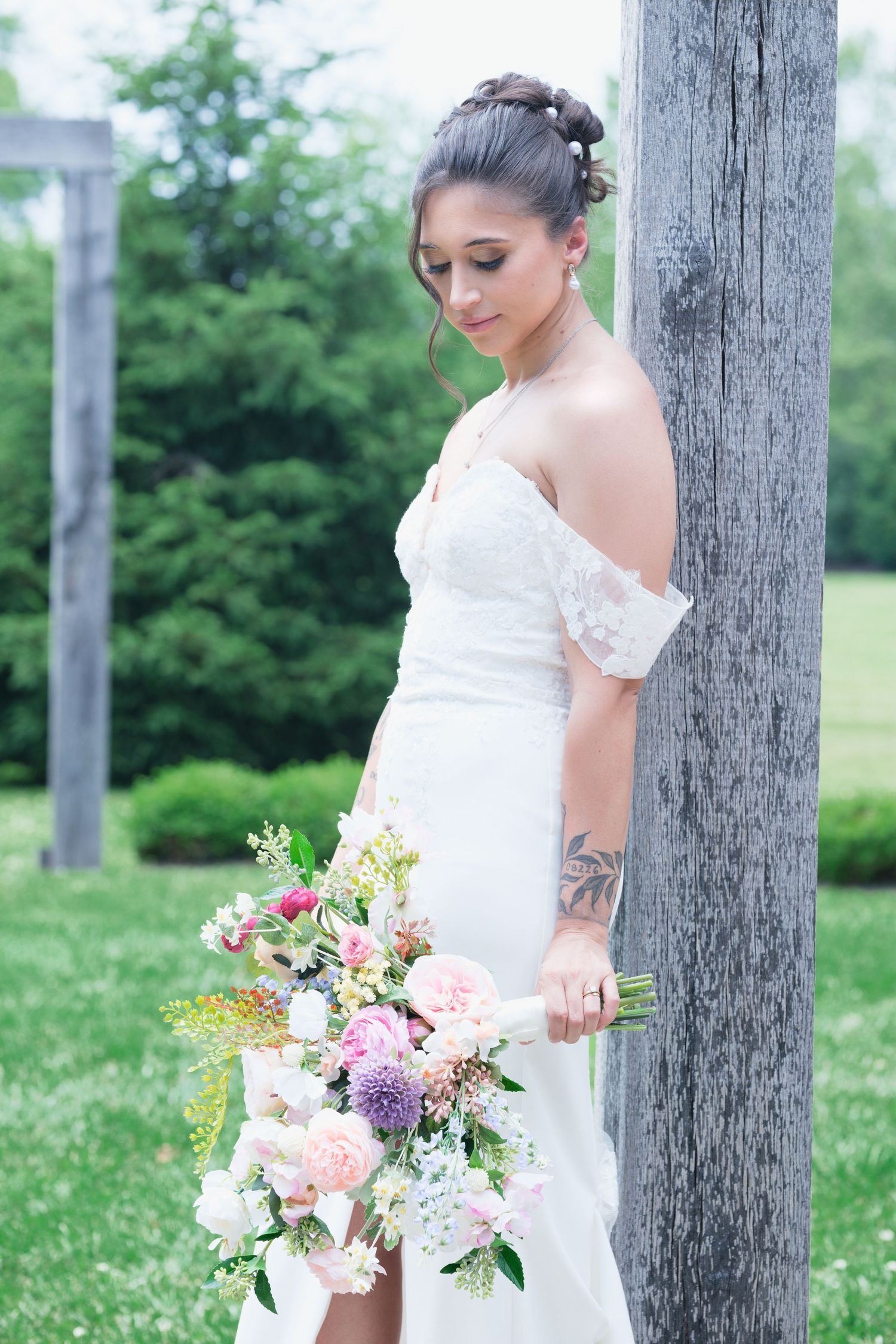 Bride in off-shoulder lace gown holding colorful bouquet of pink and purple flowers beside rustic wooden post.