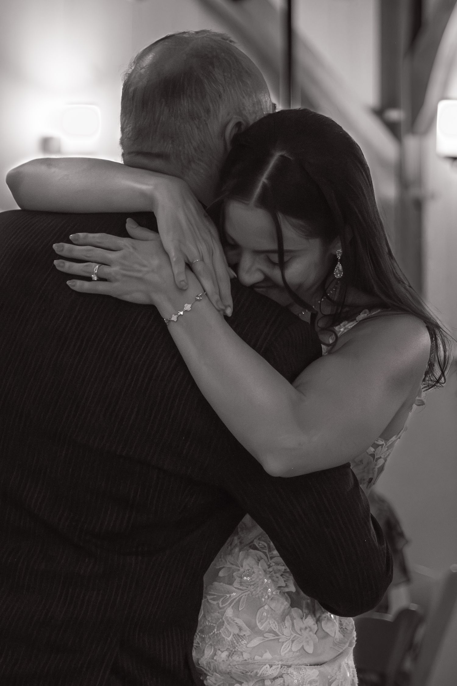 Emotional black and white photo of bride in lace dress sharing a heartfelt hug at wedding reception.
