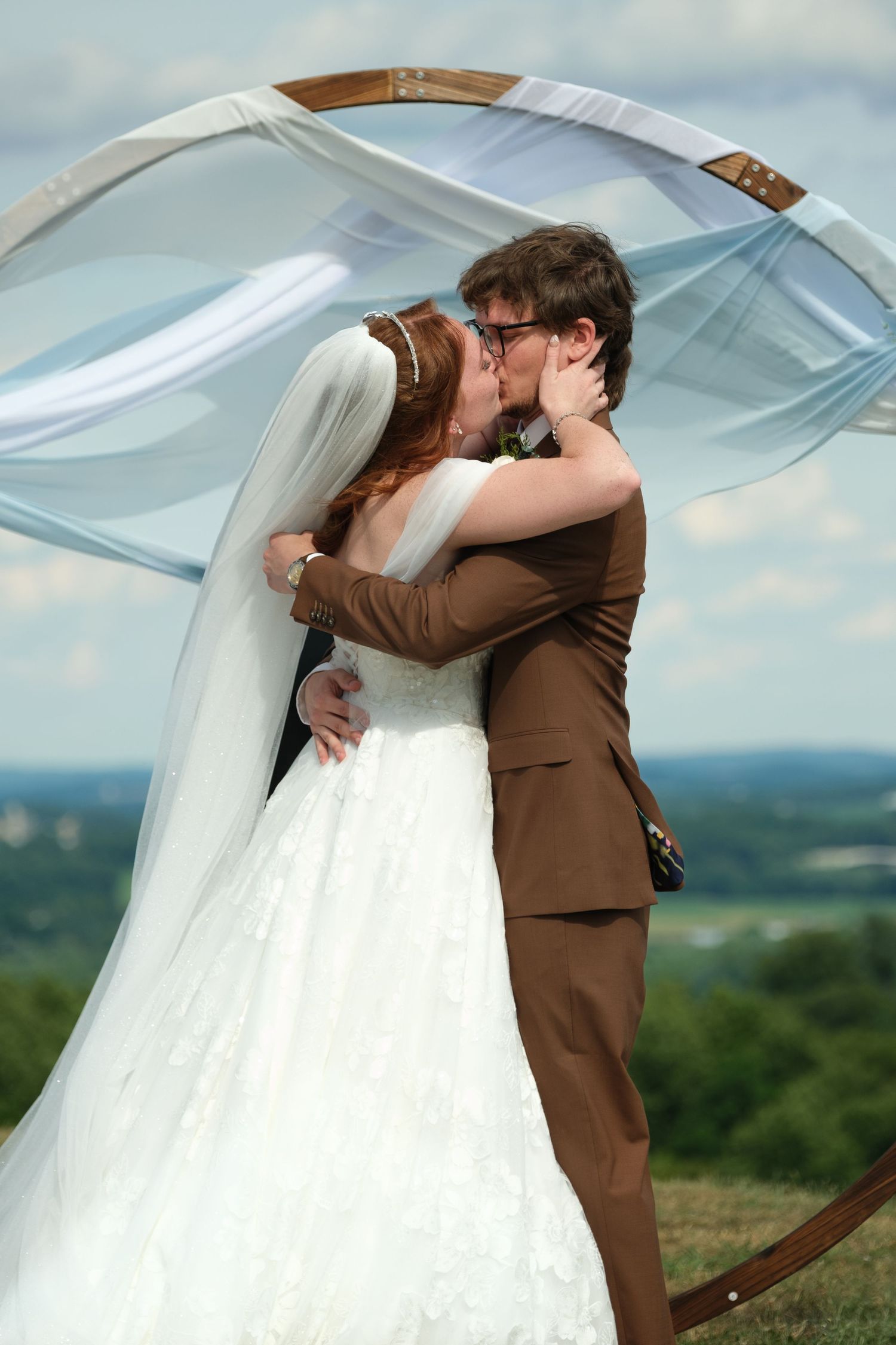 Bride and groom share first kiss under flowing arch with scenic landscape background.