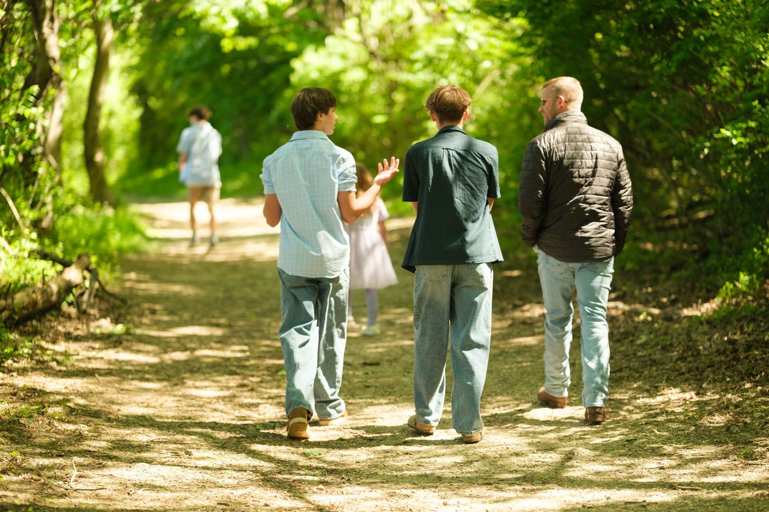 Three men walking on a sunny forest path, with two figures visible ahead among lush green trees.