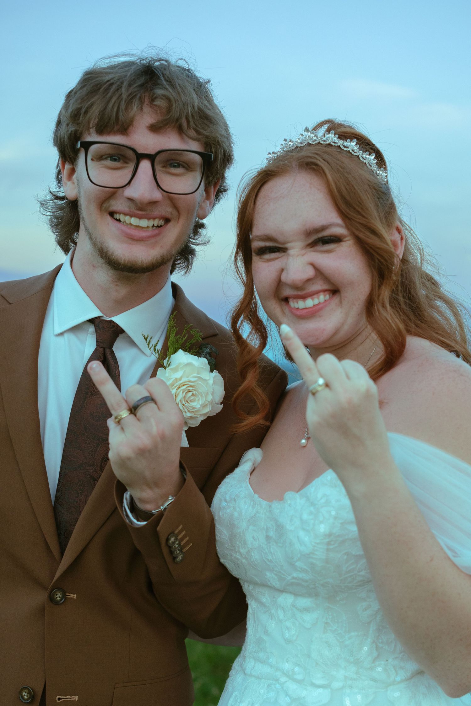 Newlyweds smiling and showing wedding rings, groom in brown suit and bride in white gown.