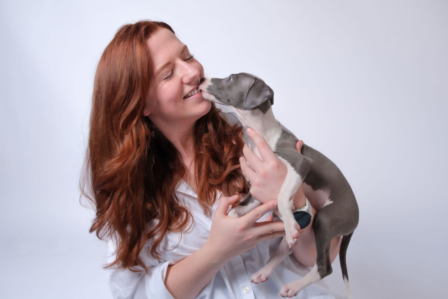 Red-haired woman in white shirt smiling as gray and white puppy licks her face on white background.