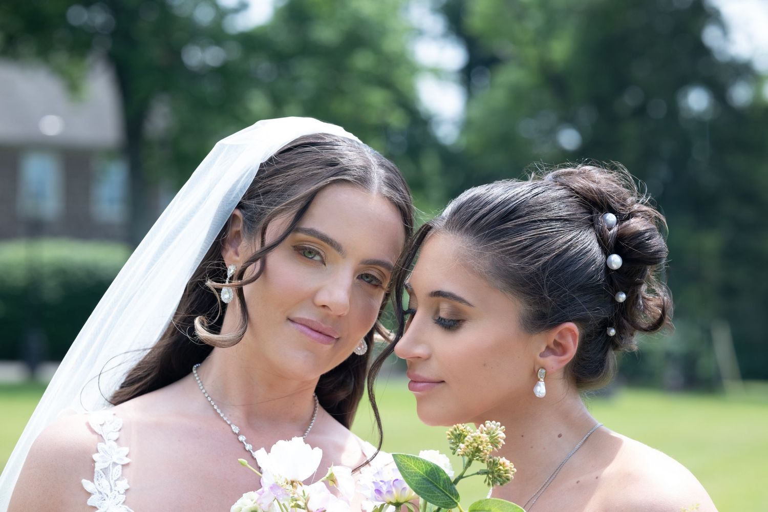 Two elegant brides in white wedding attire with veils and pearl accessories pose together outdoors at a garden wedding.