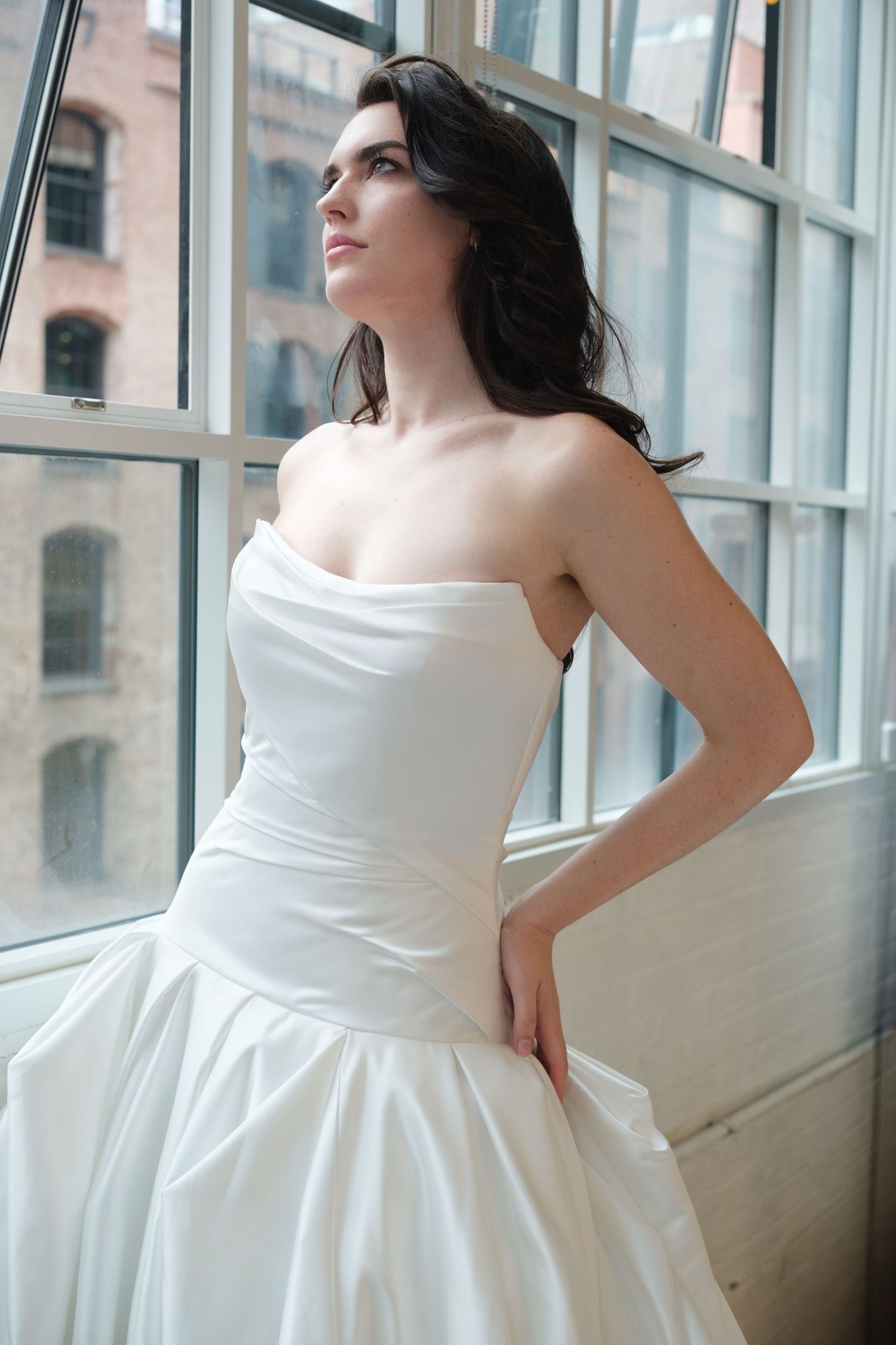 Elegant bride in a strapless white satin ball gown gazing out large windows in a loft setting.