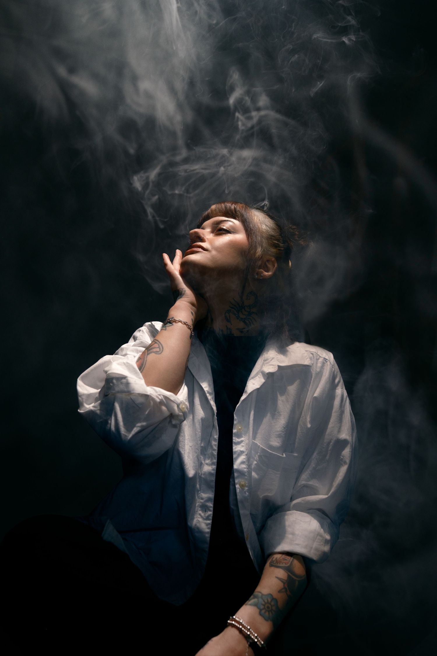 A tattooed woman in a white shirt poses dramatically surrounded by swirling smoke against a dark background.