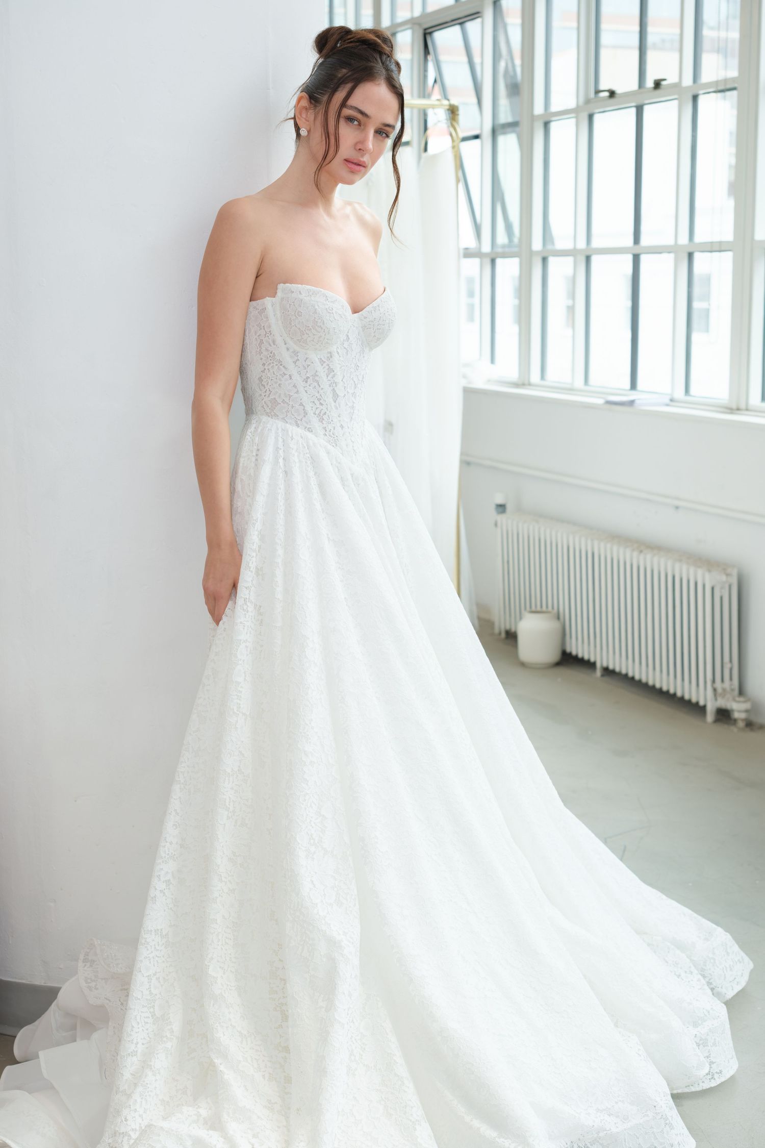 Bride in a strapless white lace wedding ball gown with a corseted sweetheart bodice and full skirt, posed beside a bright industrial window.