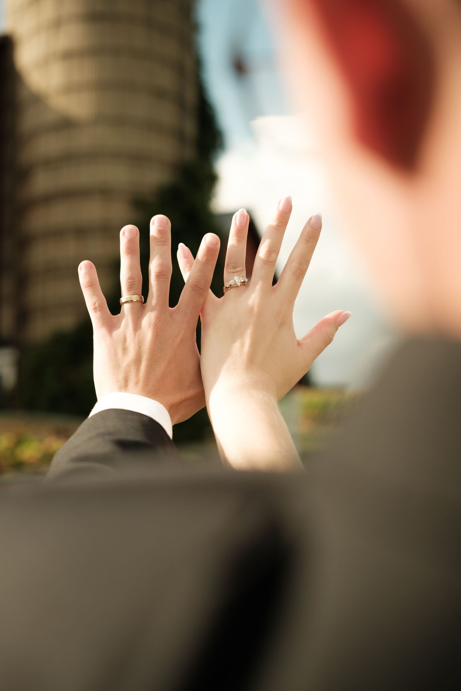 Newlywed couple holding up their hands side by side to show wedding rings, with a tall silo-like building blurred in the background.