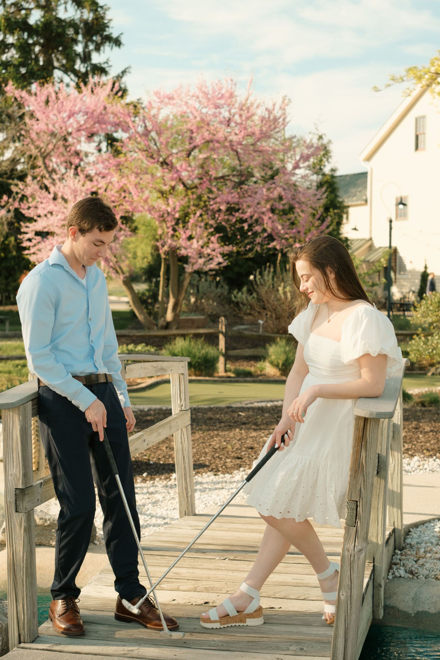 A young couple plays mini golf on a wooden bridge surrounded by blooming pink trees in spring.