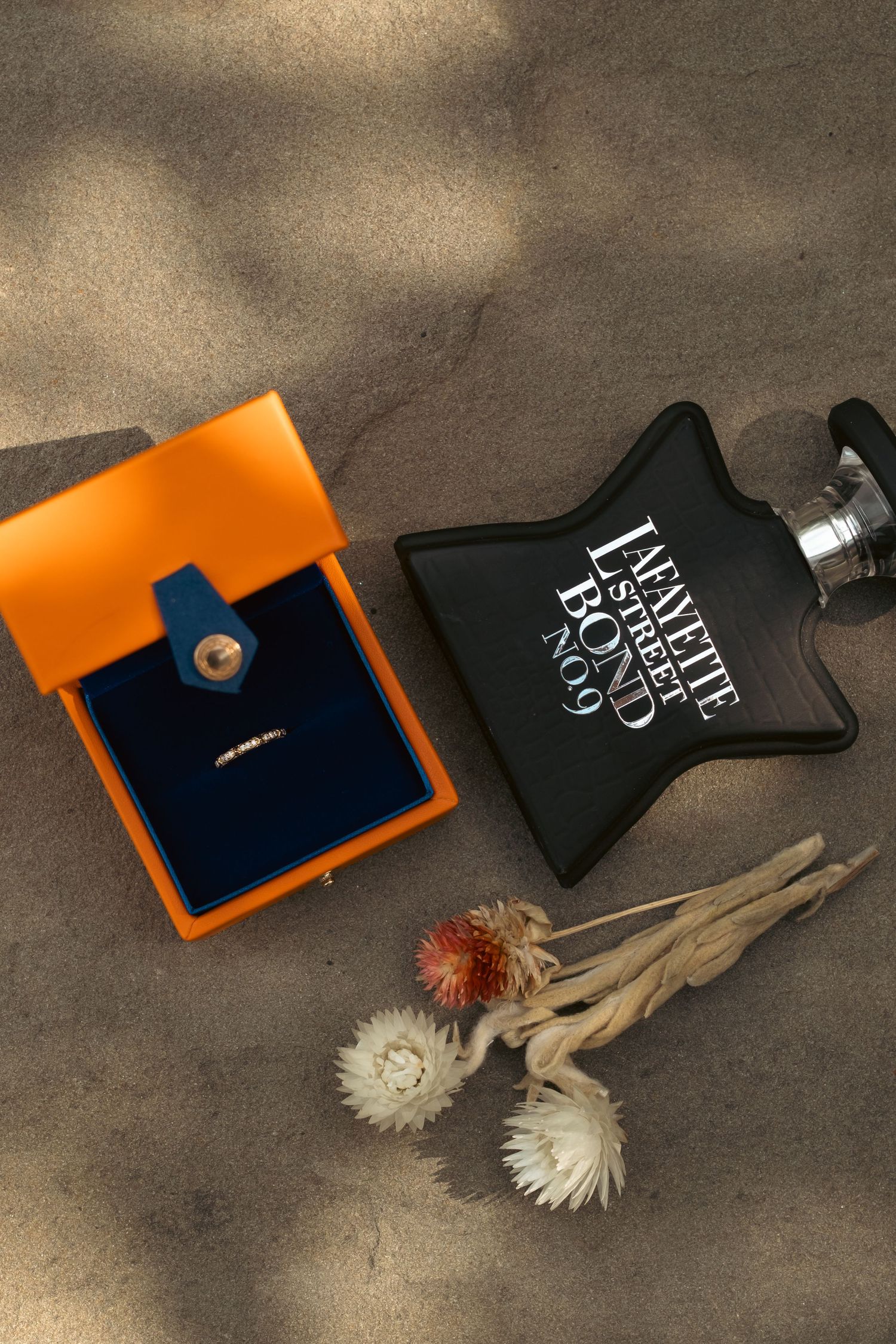 Bond No. 9 Lafayette Street perfume bottle beside an orange jewelry box and dried flowers on sandy surface.