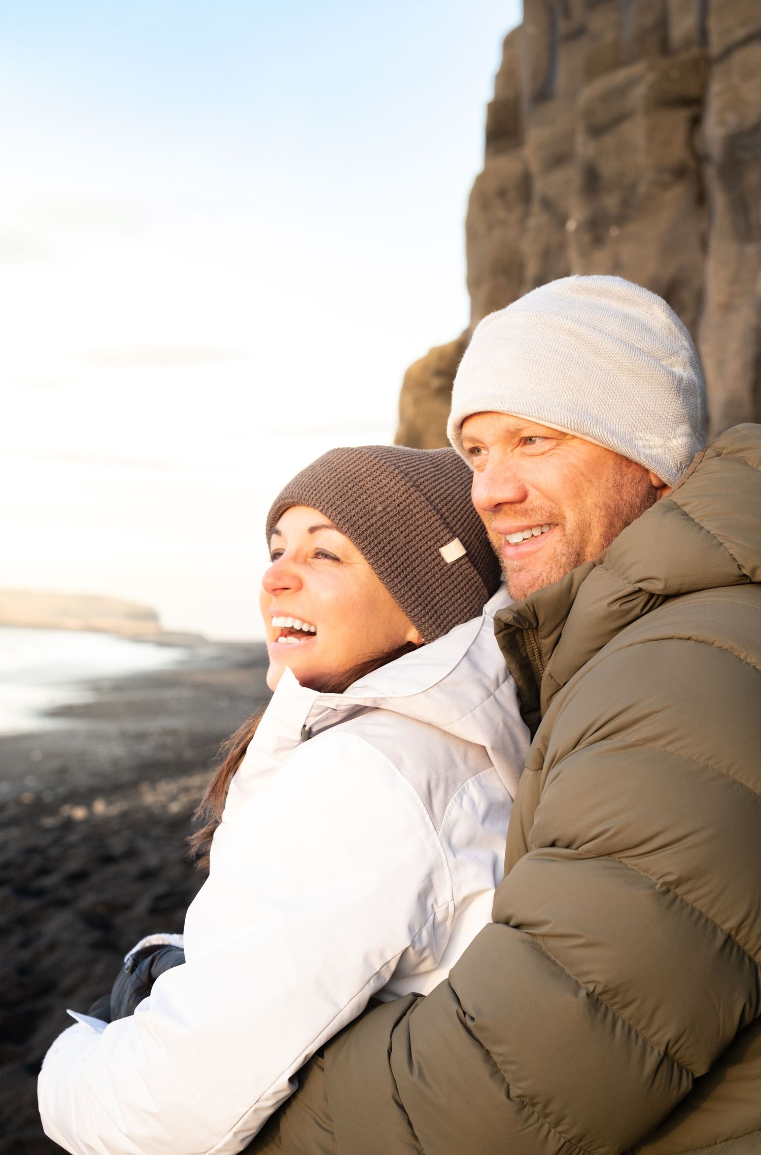 Couple in winter coats and knit hats embrace outdoors during golden hour.