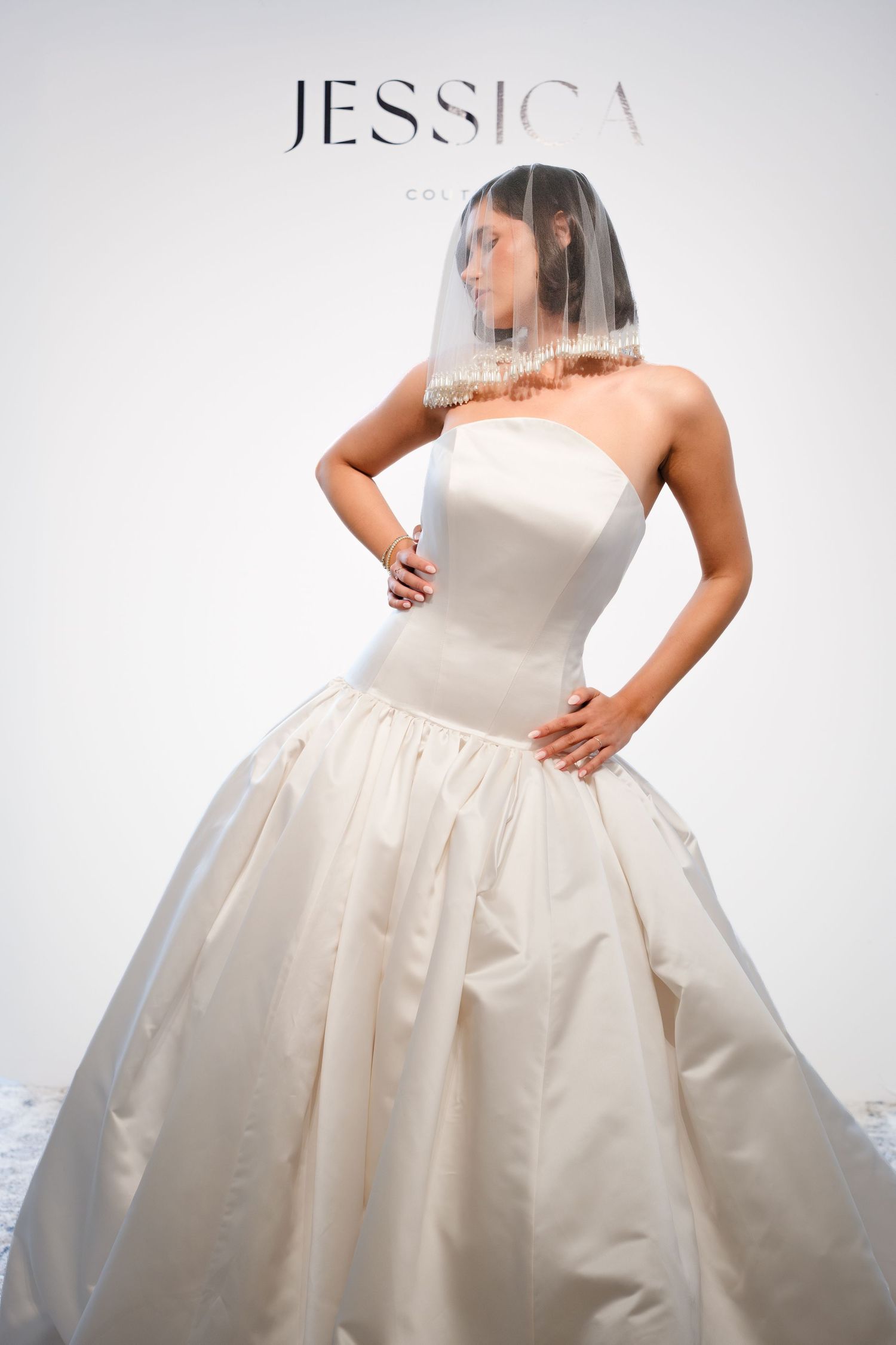Bride in a strapless ivory satin ball gown with a full pleated skirt and a sheer blusher veil over her face, posed against a white backdrop.