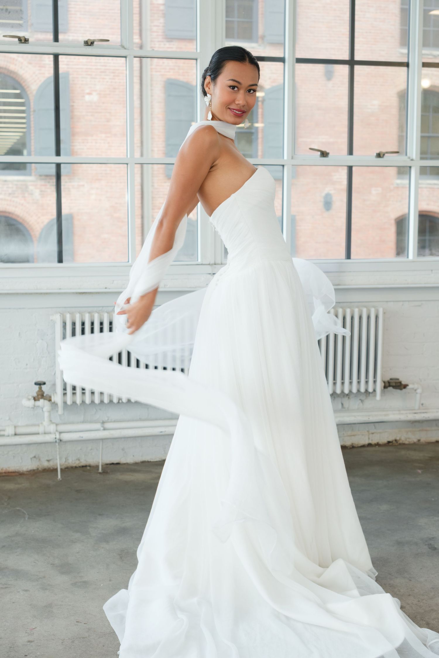 A bride in a flowing white one-shoulder wedding gown twirls gracefully in a bright industrial loft space.