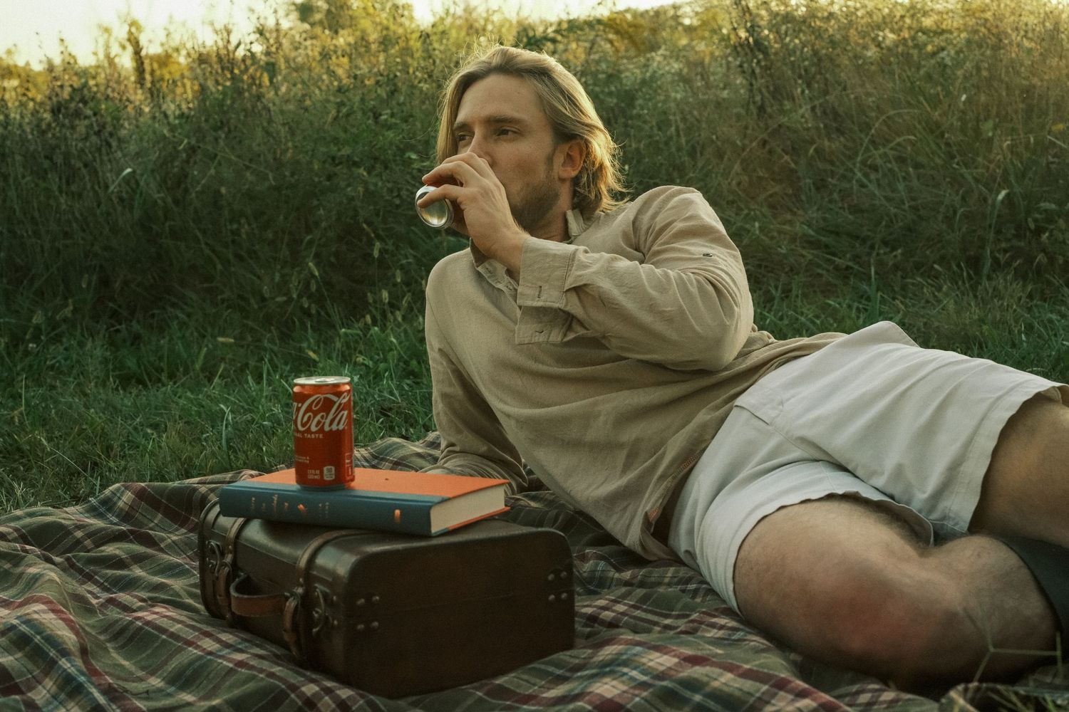 Man relaxing on a picnic blanket outdoors, drinking from a glass beside a Coca-Cola can and books.
