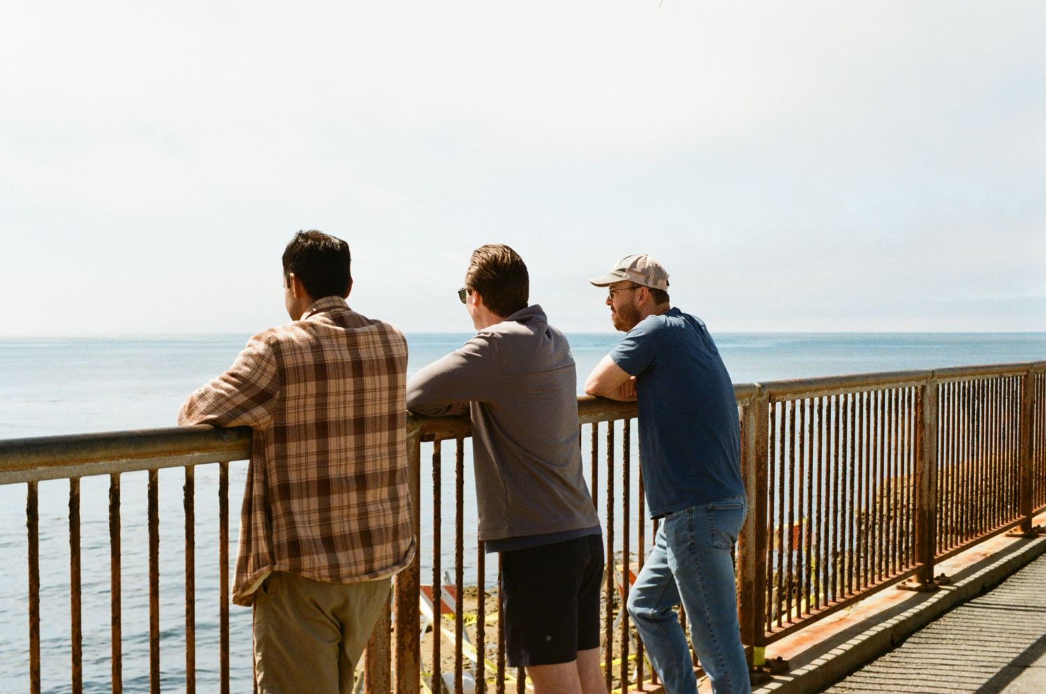 Three men lean on a wooden railing overlooking a calm ocean horizon on a clear day.