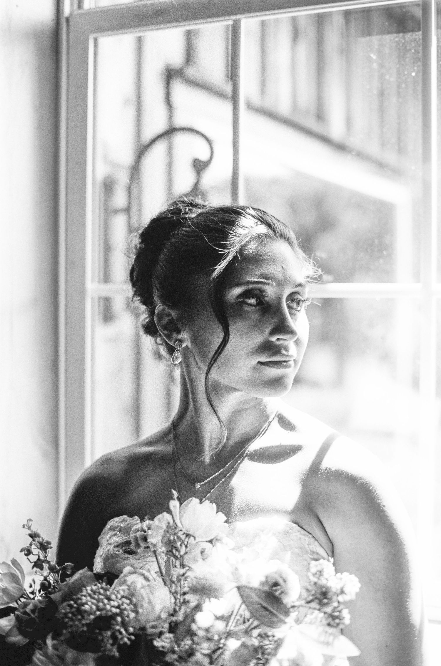 Black and white portrait of a bride gazing out a window while holding a bouquet of fresh flowers.