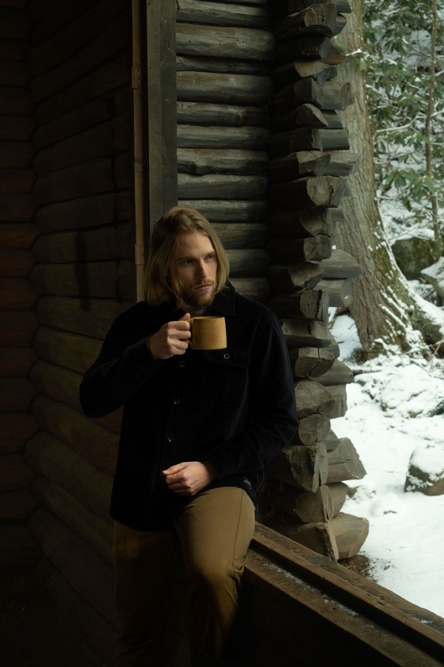 Someone drinks from a mug while sitting on a rustic log cabin porch in winter with snow visible outside.