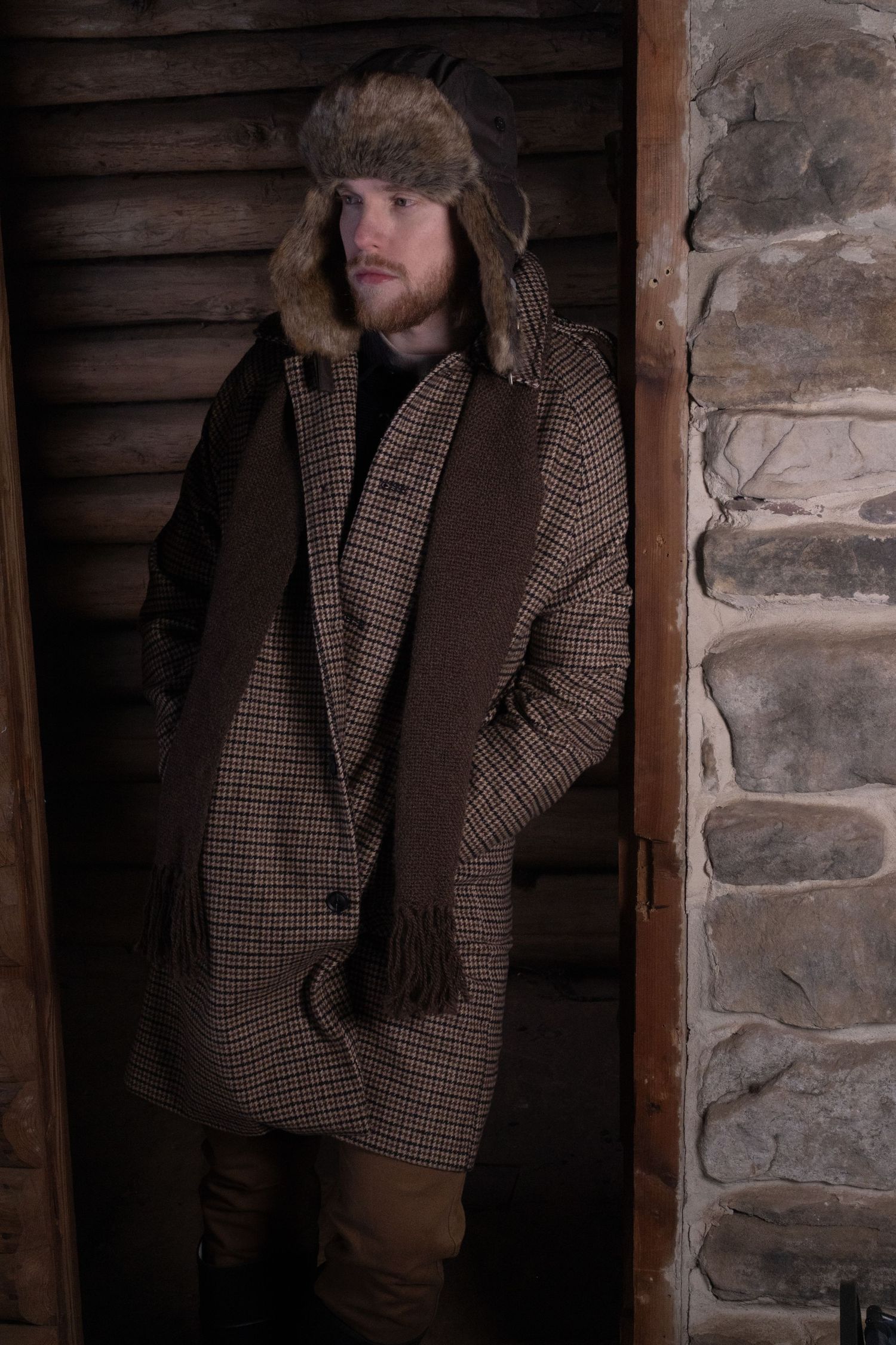 Person wearing brown tweed coat and cap stands against rustic log cabin wall in moody lighting.
