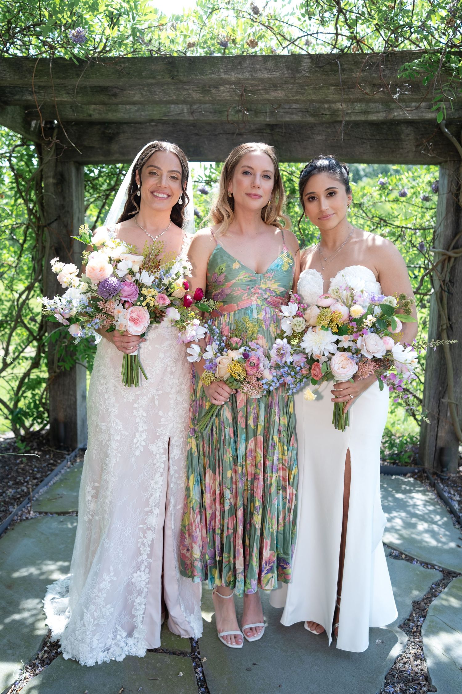 Three women in wedding attire holding floral bouquets posing under a wooden garden arbor with greenery.
