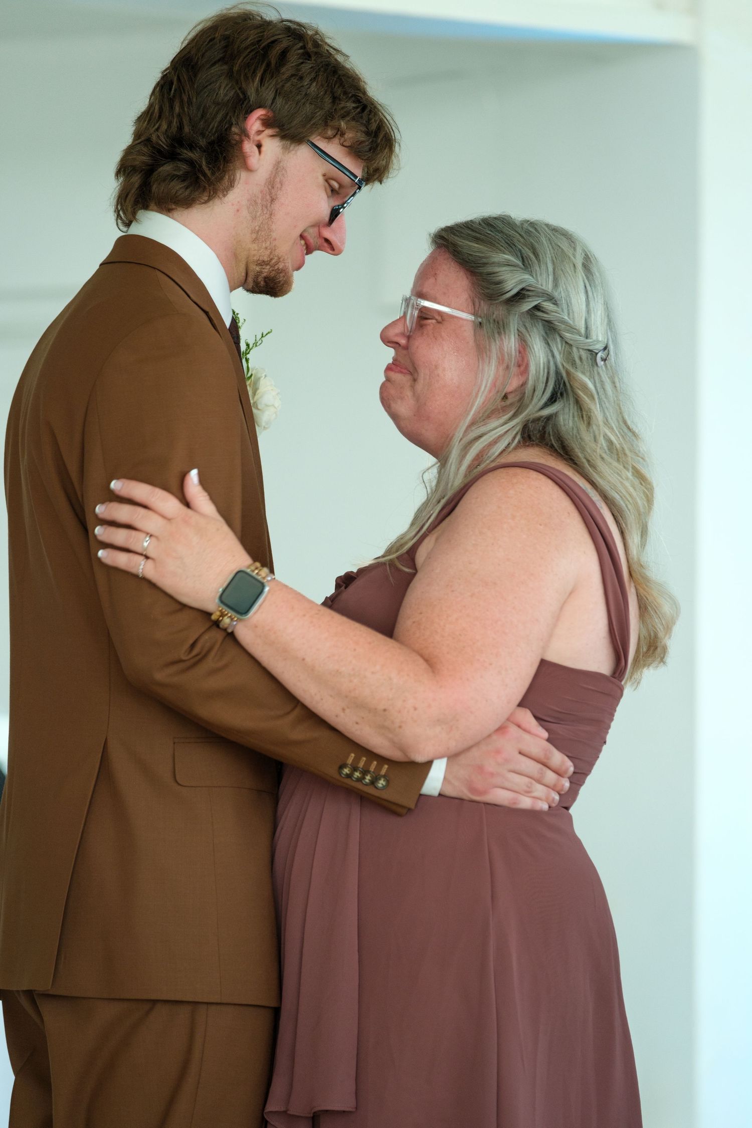 A groom in brown suit shares an emotional dance with a woman in mauve dress at a wedding reception.