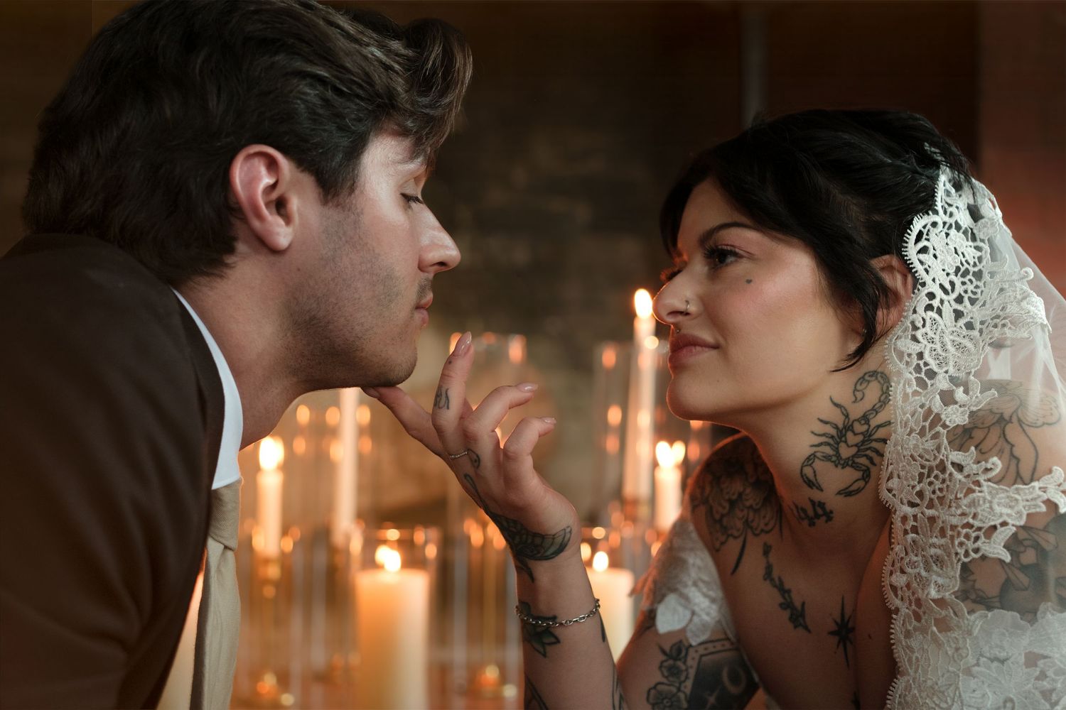 Romantic couple sharing an intimate moment surrounded by candlelight, bride in lace veil with tattoos gazing at partner.