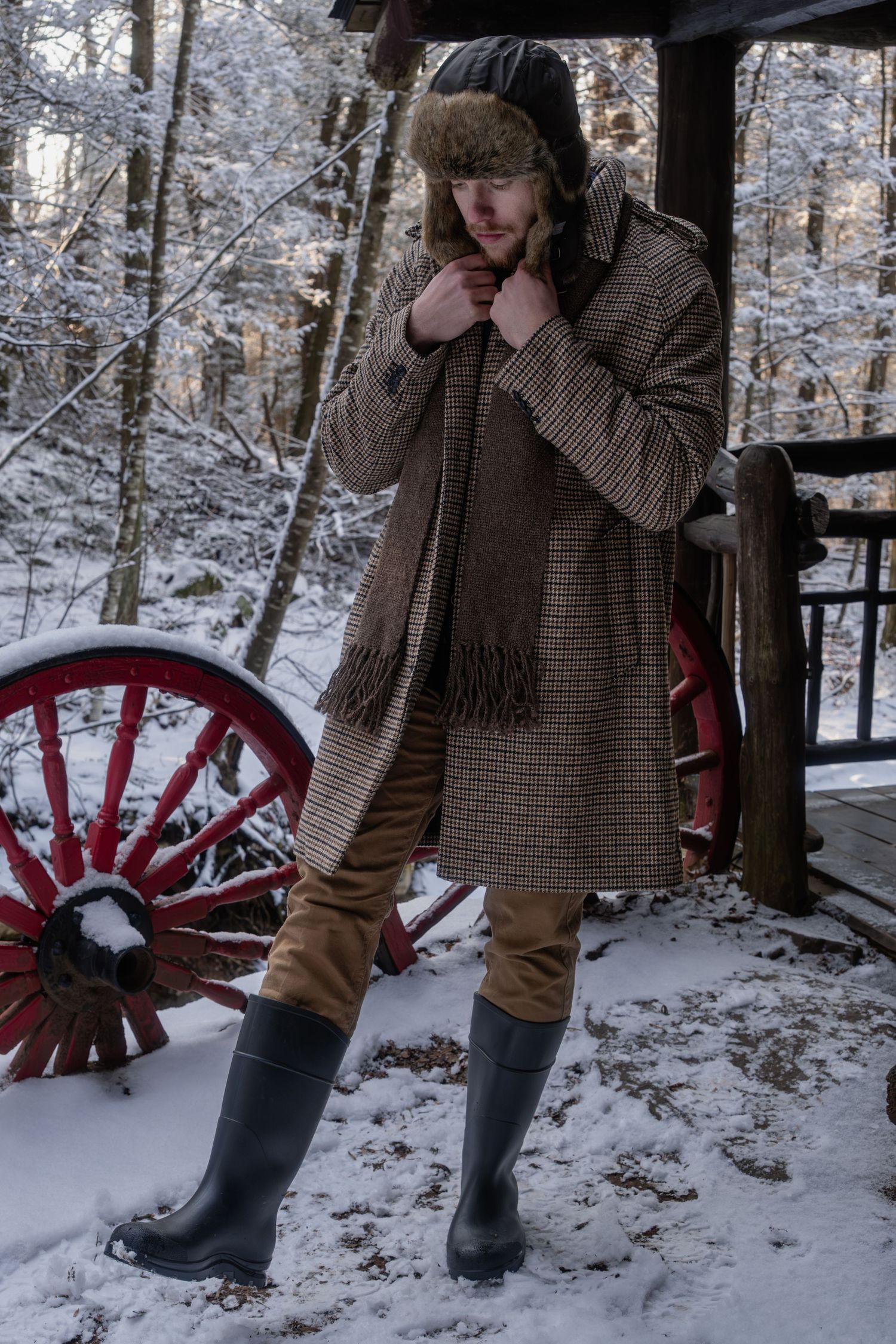 Someone in winter attire with a fur hat and boots stands on a snowy porch with a red wagon wheel in the background.