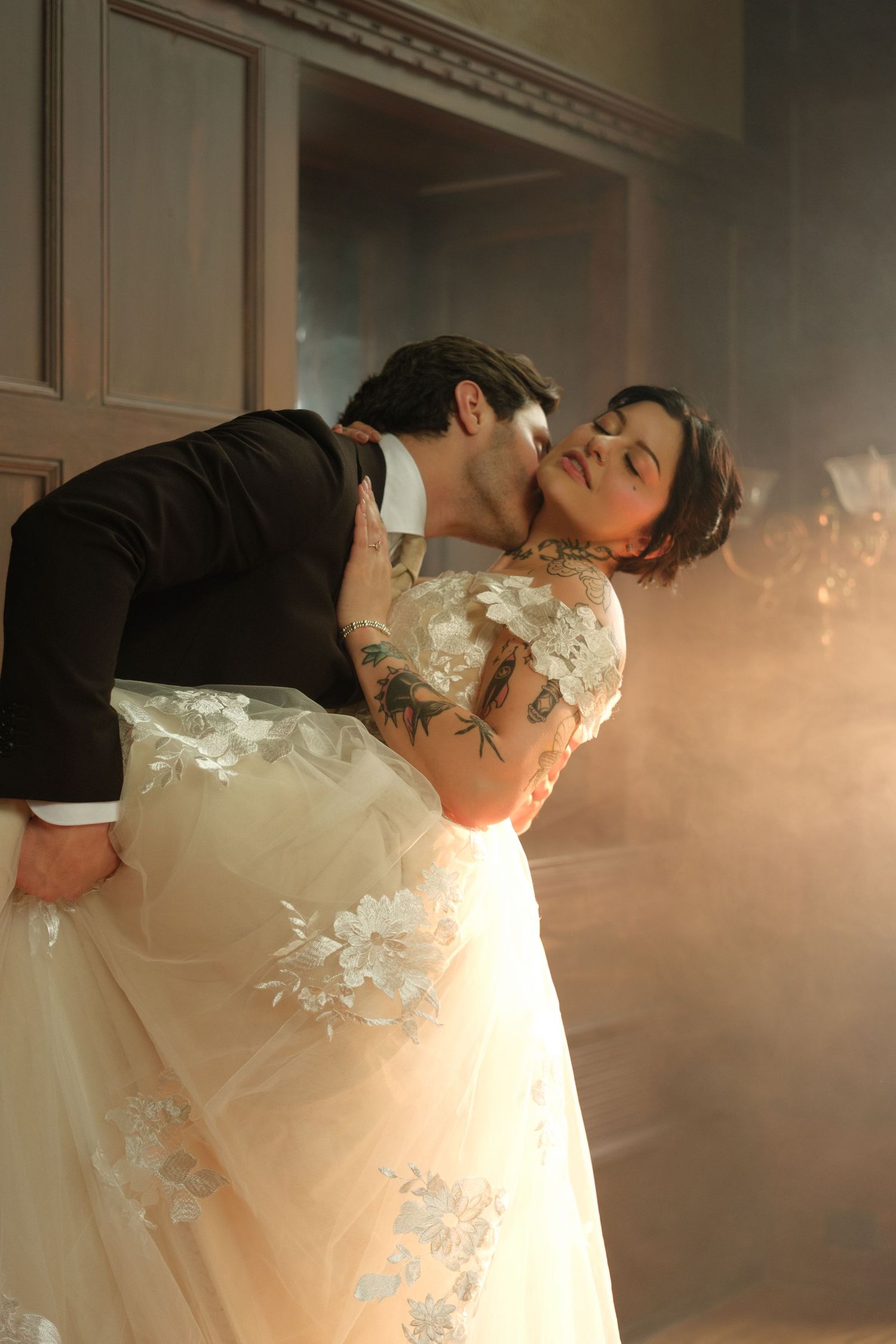 Bride in floral gown dips romantically with groom in black suit, surrounded by dreamy smoke at wedding.