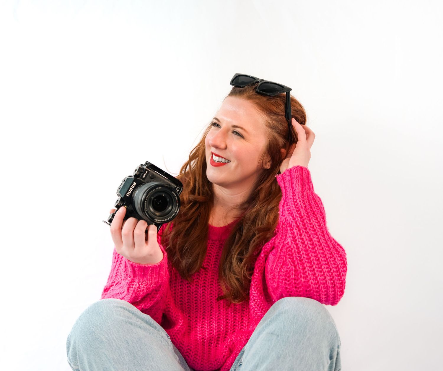 Photographer in bright pink sweater laughs while holding DSLR camera against white background.