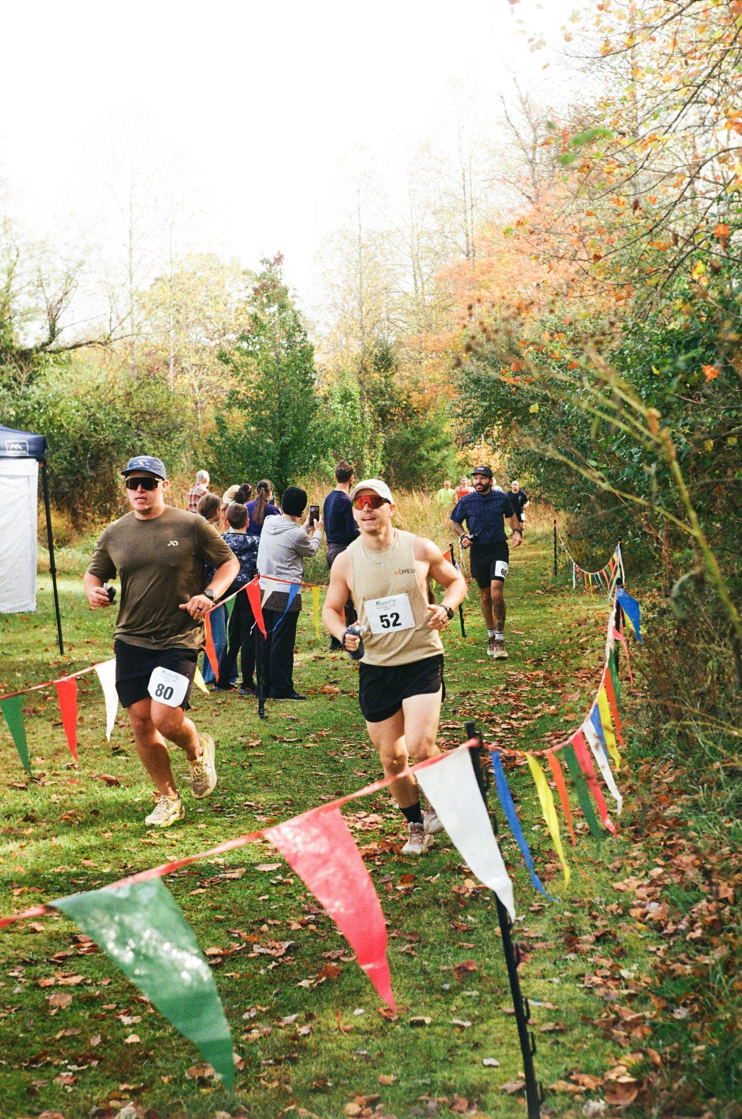 Runners navigate through a wooded trail course marked with colorful flags during an autumn cross-country race event.