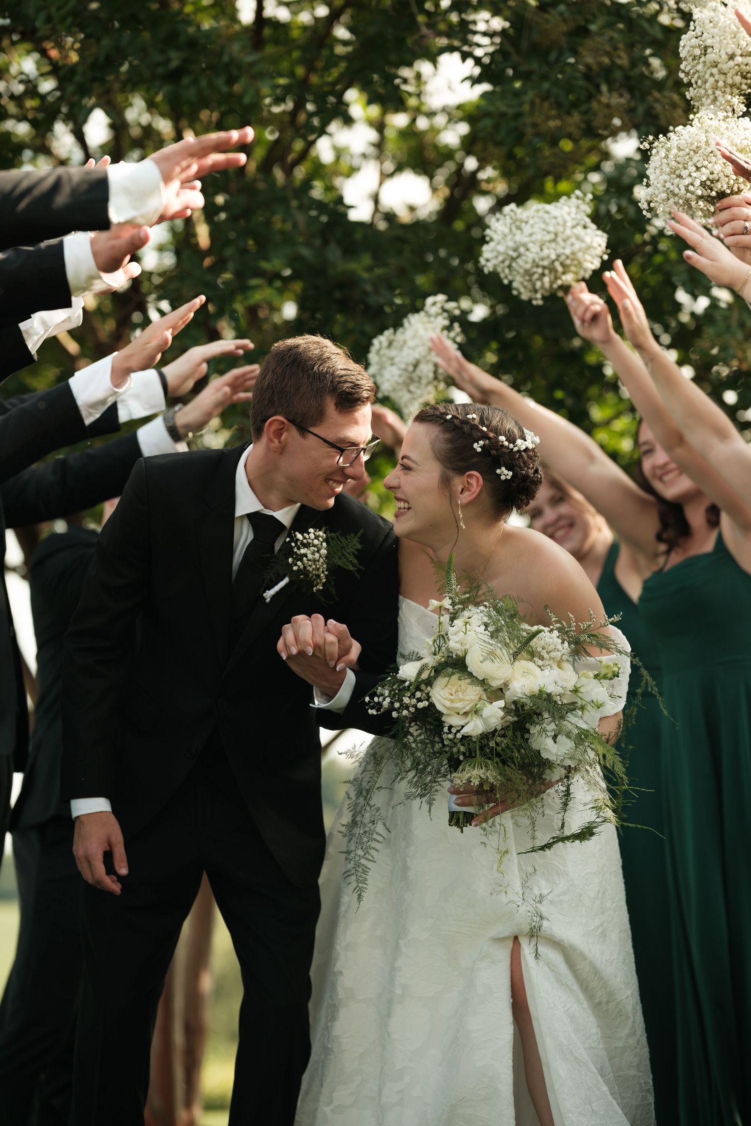 Bride and groom hold hands smiling, surrounded by wedding party raising bouquets outdoors.