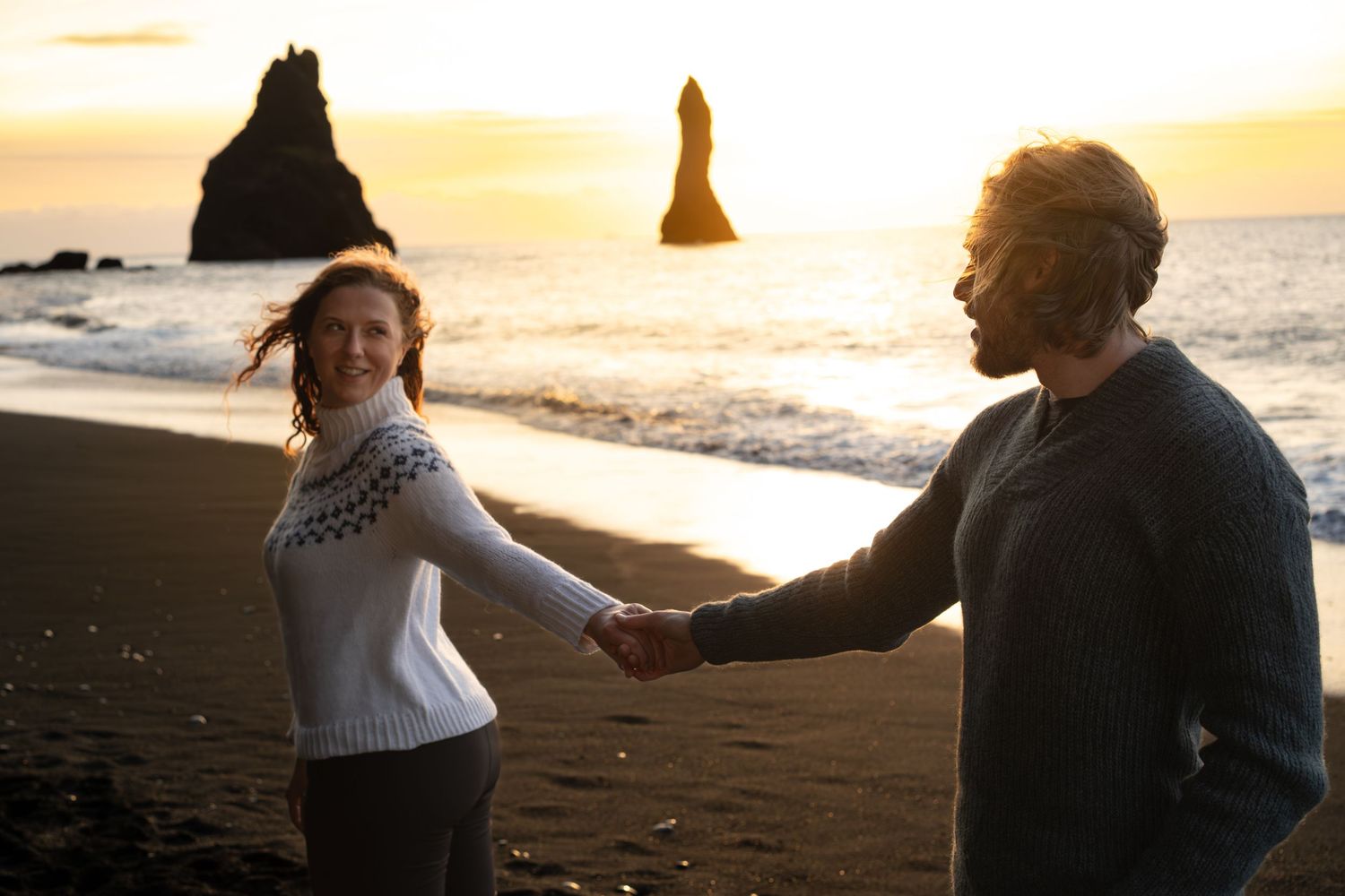 A couple holds hands and walks along a scenic beach at sunset with warm golden light illuminating their silhouettes.