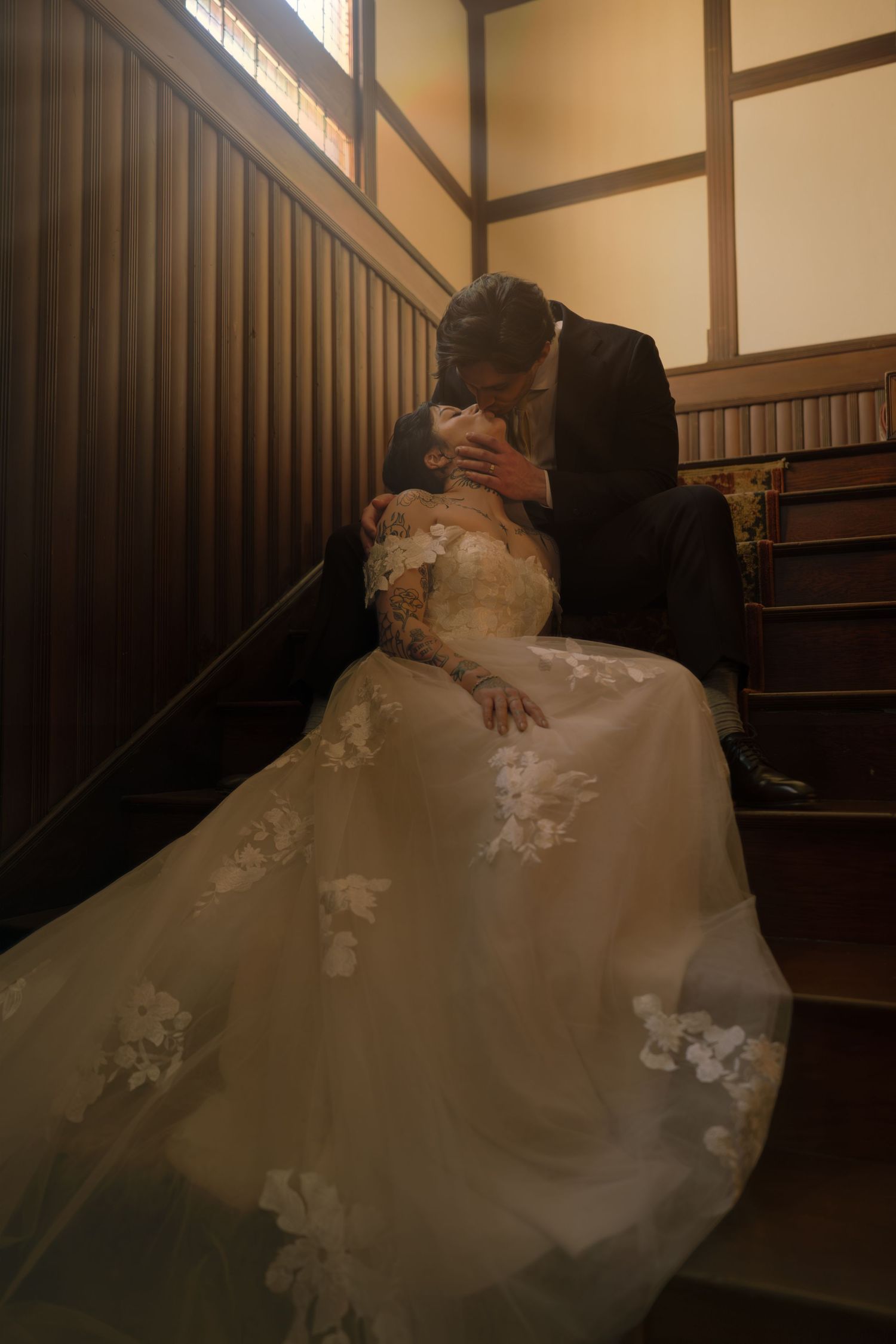 Bride and groom share a romantic kiss on wooden staircase, bride wearing elegant floral lace wedding gown.