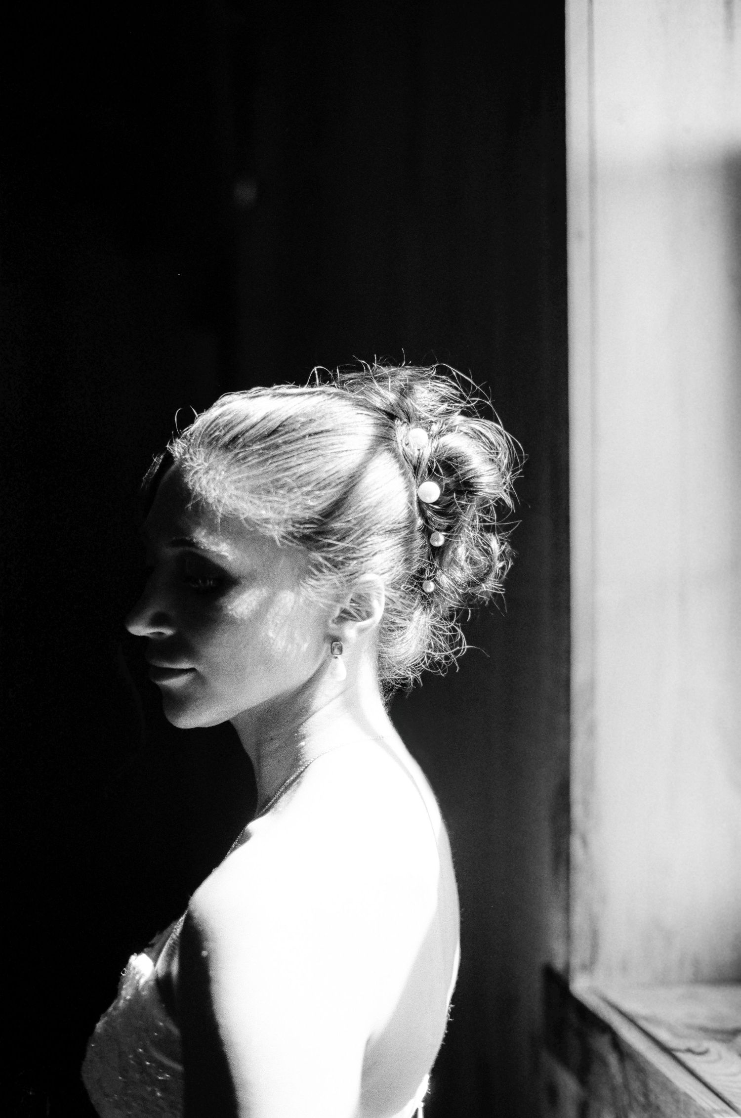 Black and white profile portrait of a bride with elegant updo hairstyle and pearl hair accessories by window.