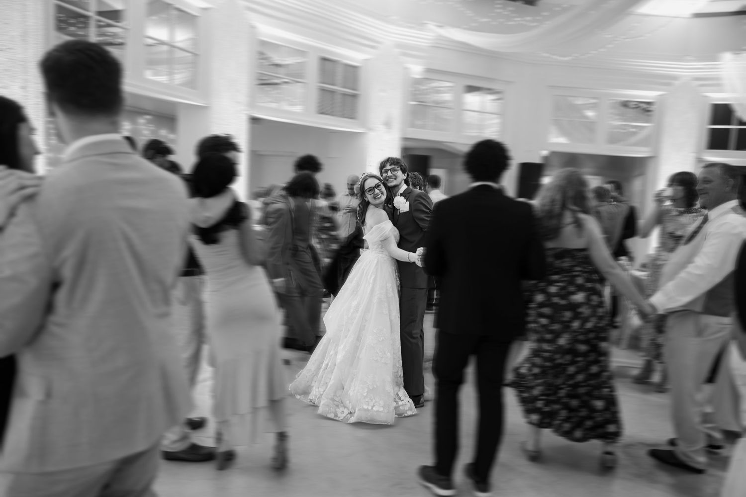 Black and white photo of bride and groom sharing first dance surrounded by guests at elegant wedding reception.