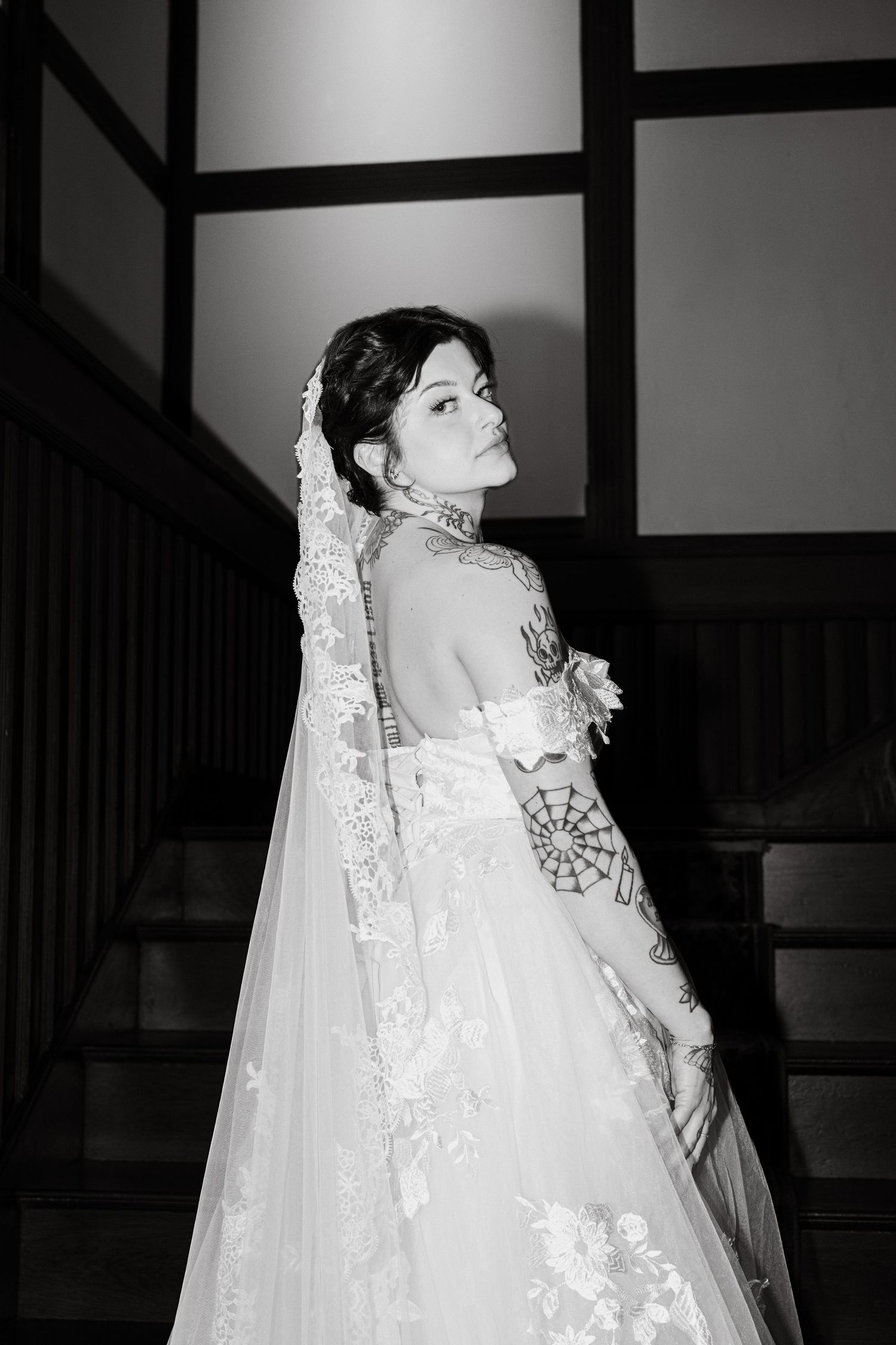 Black and white photo of tattooed bride in lace wedding gown and veil, looking over shoulder on staircase.