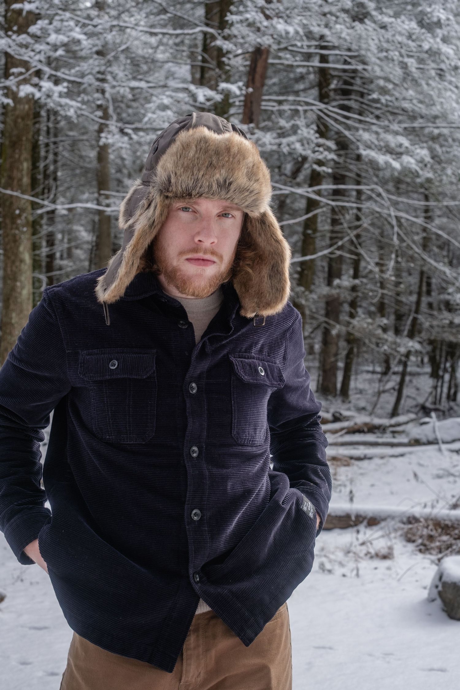 A person in a fur hat and dark jacket smiles while walking through a snowy winter forest landscape.