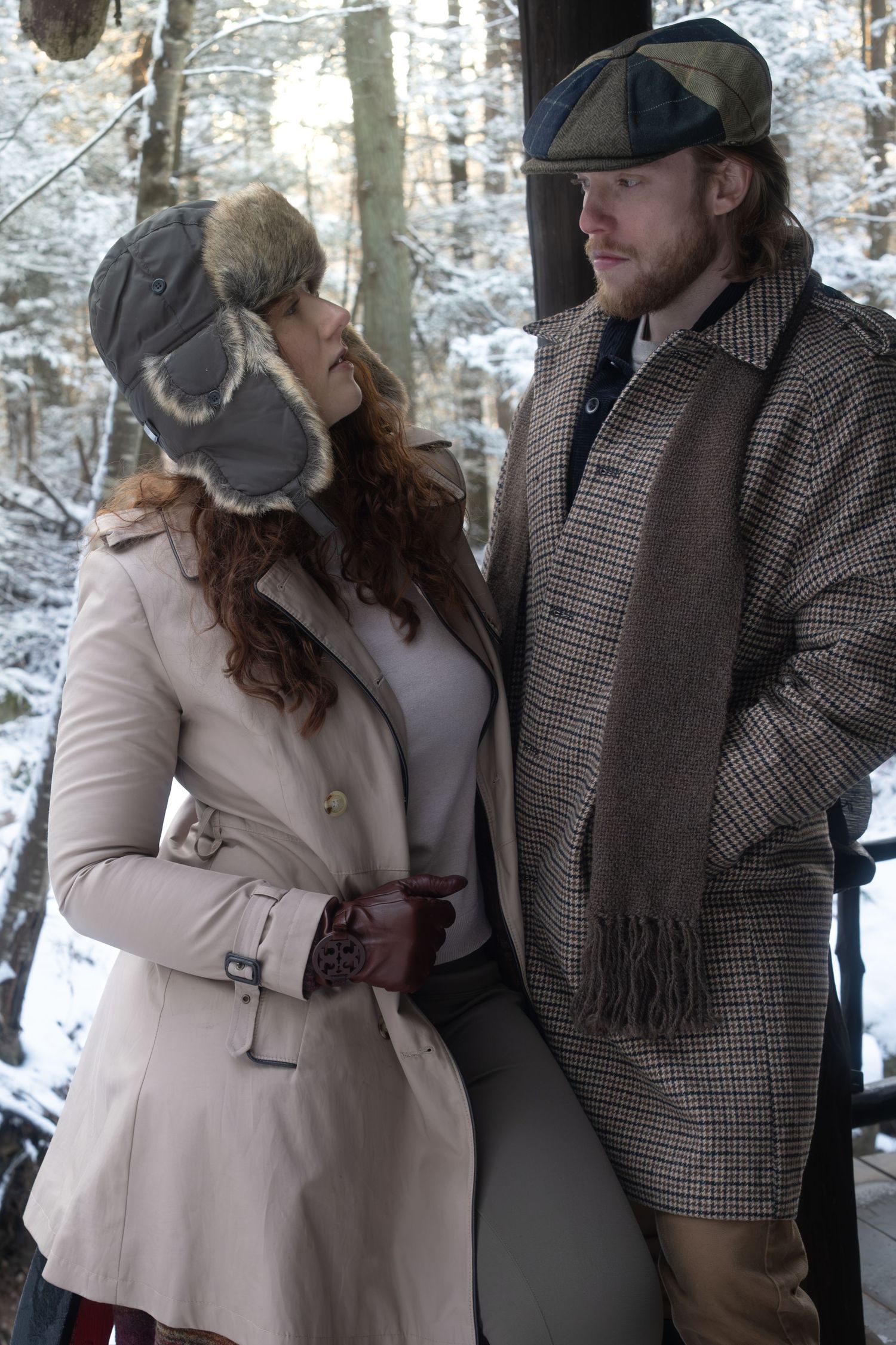 A couple in winter coats and hats share an intimate moment in a snowy forest setting.