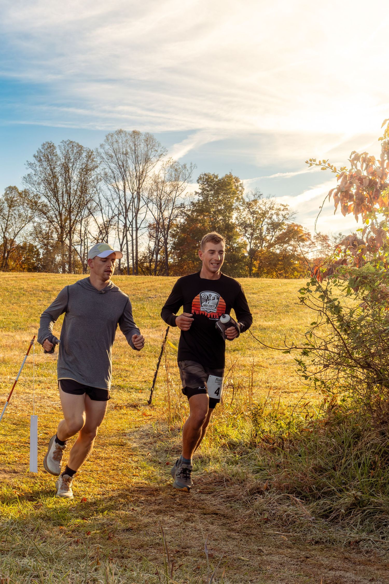 Two runners jog together on a dirt trail during golden hour with autumn trees and sunlight in the background.