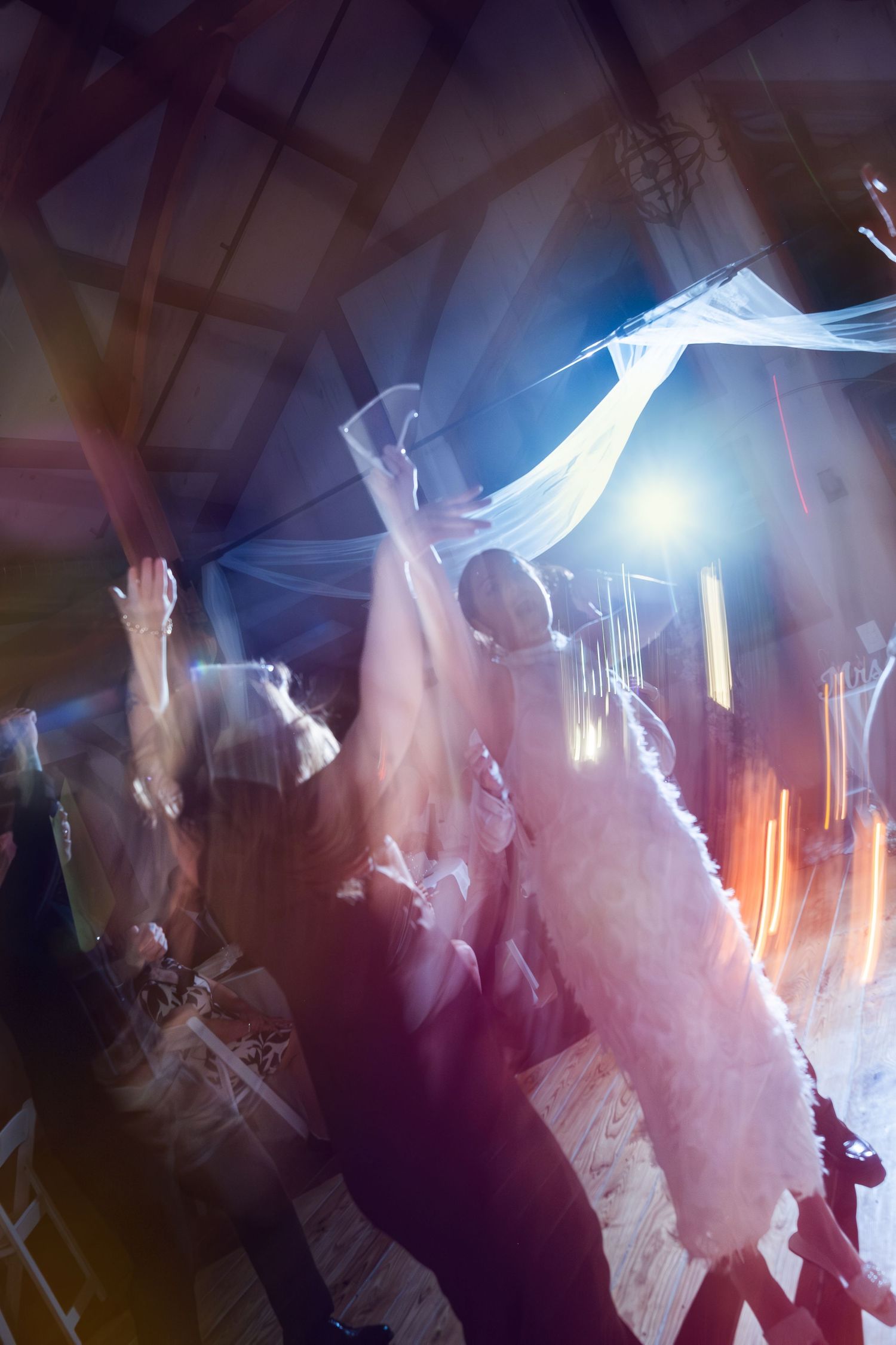 Bride dancing joyfully with guests at a rustic barn wedding reception, veil flowing under colorful lights.