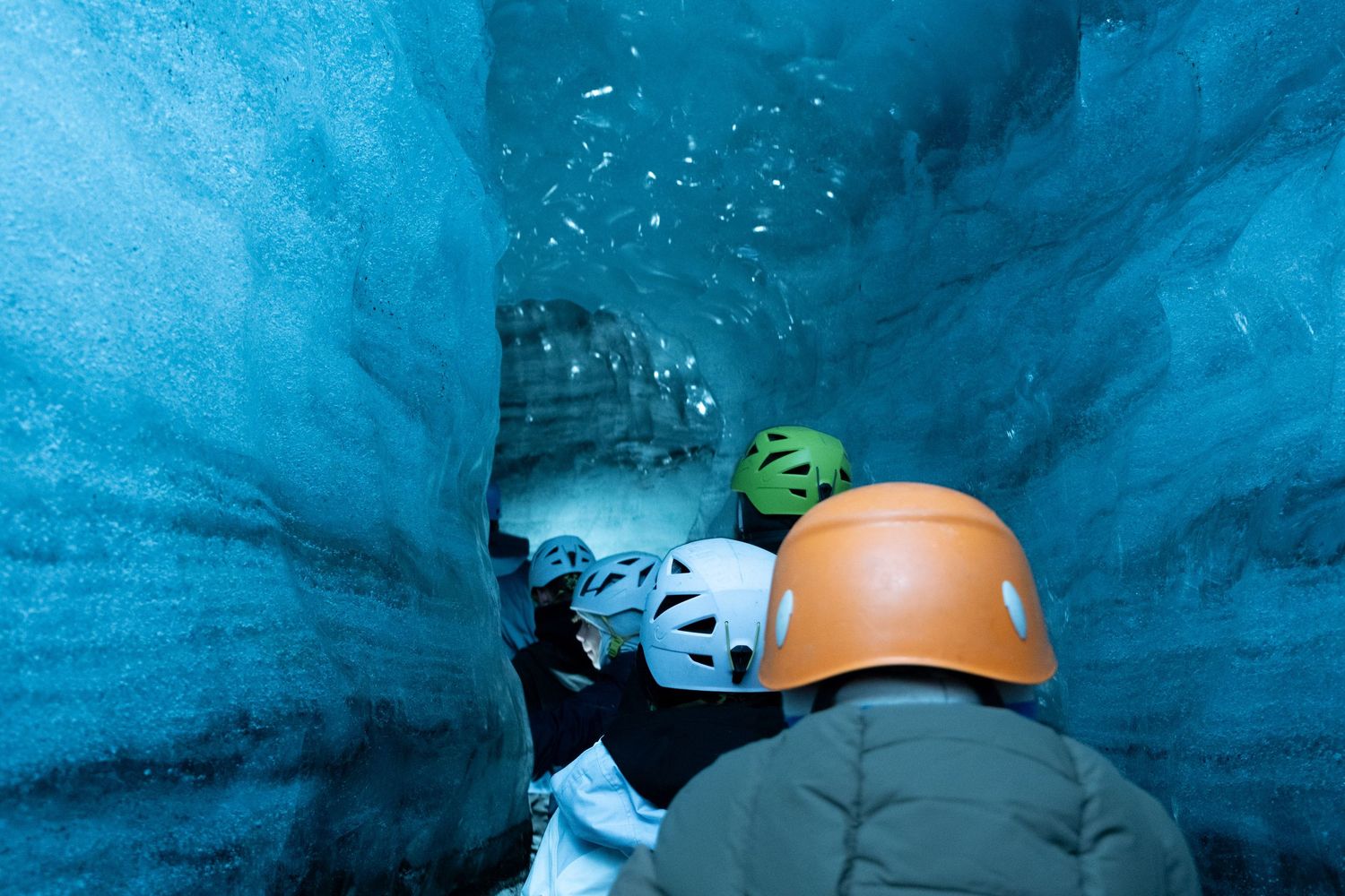 Tourists wearing safety helmets explore inside a bright blue ice cave in a glacier.