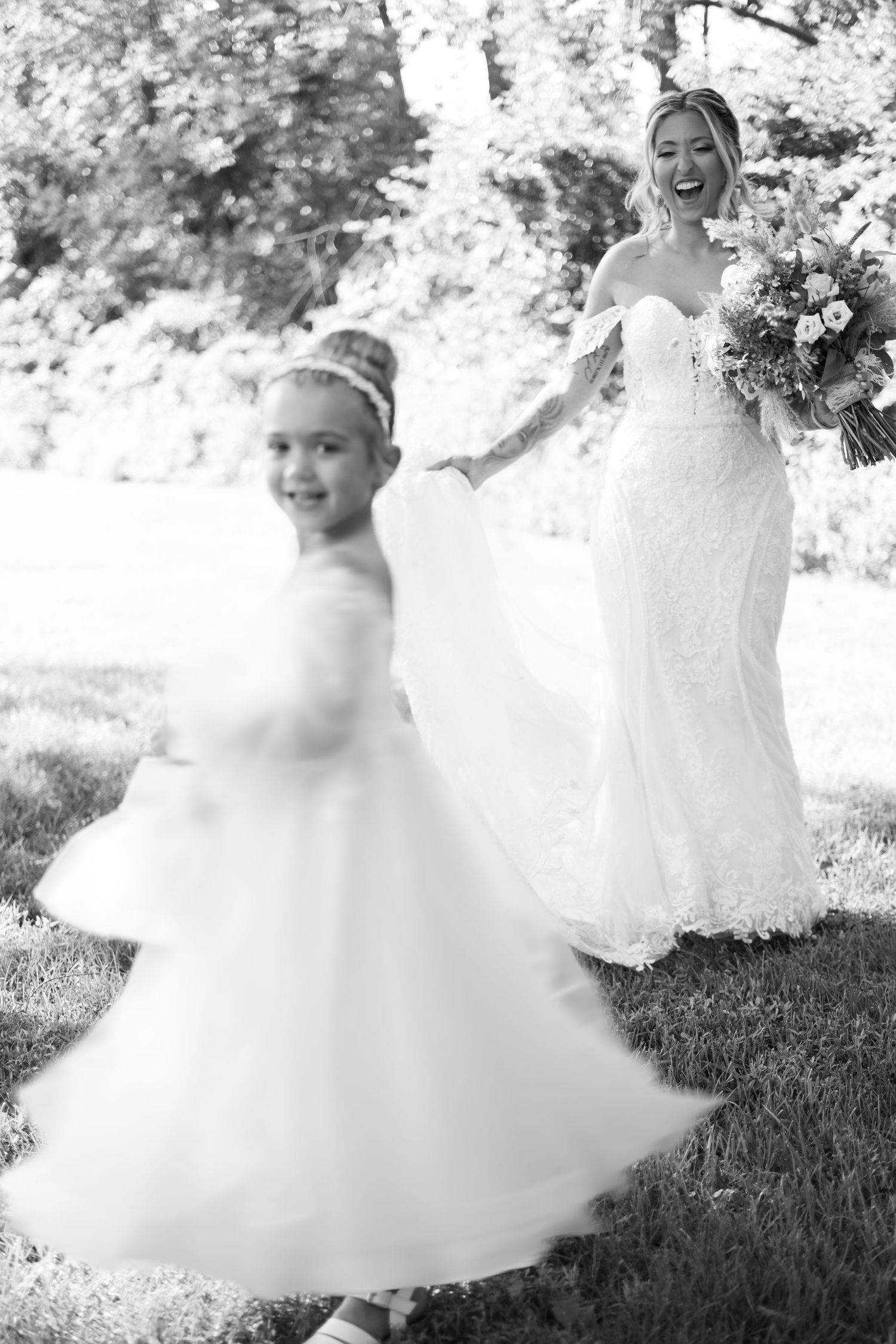 Black and white photo of a smiling bride in a lace gown holding a bouquet as a flower girl twirls in a tulle dress on a sunny lawn.
