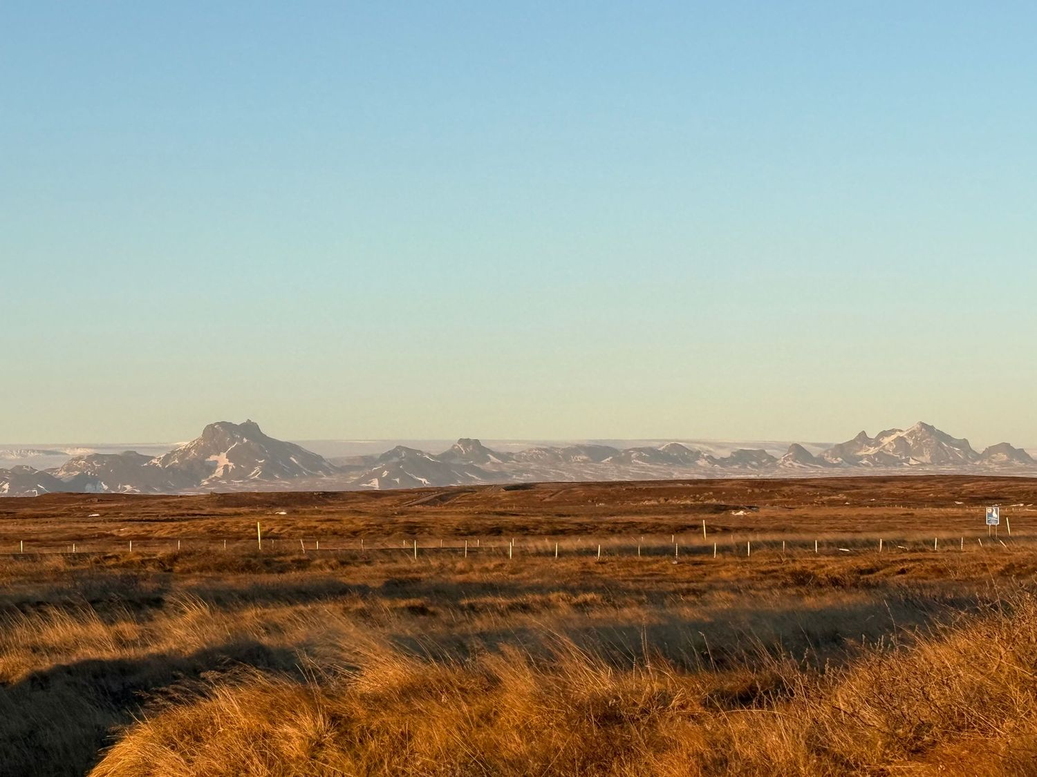 Golden prairie grass stretches toward distant mountains under clear blue sky in rural landscape.