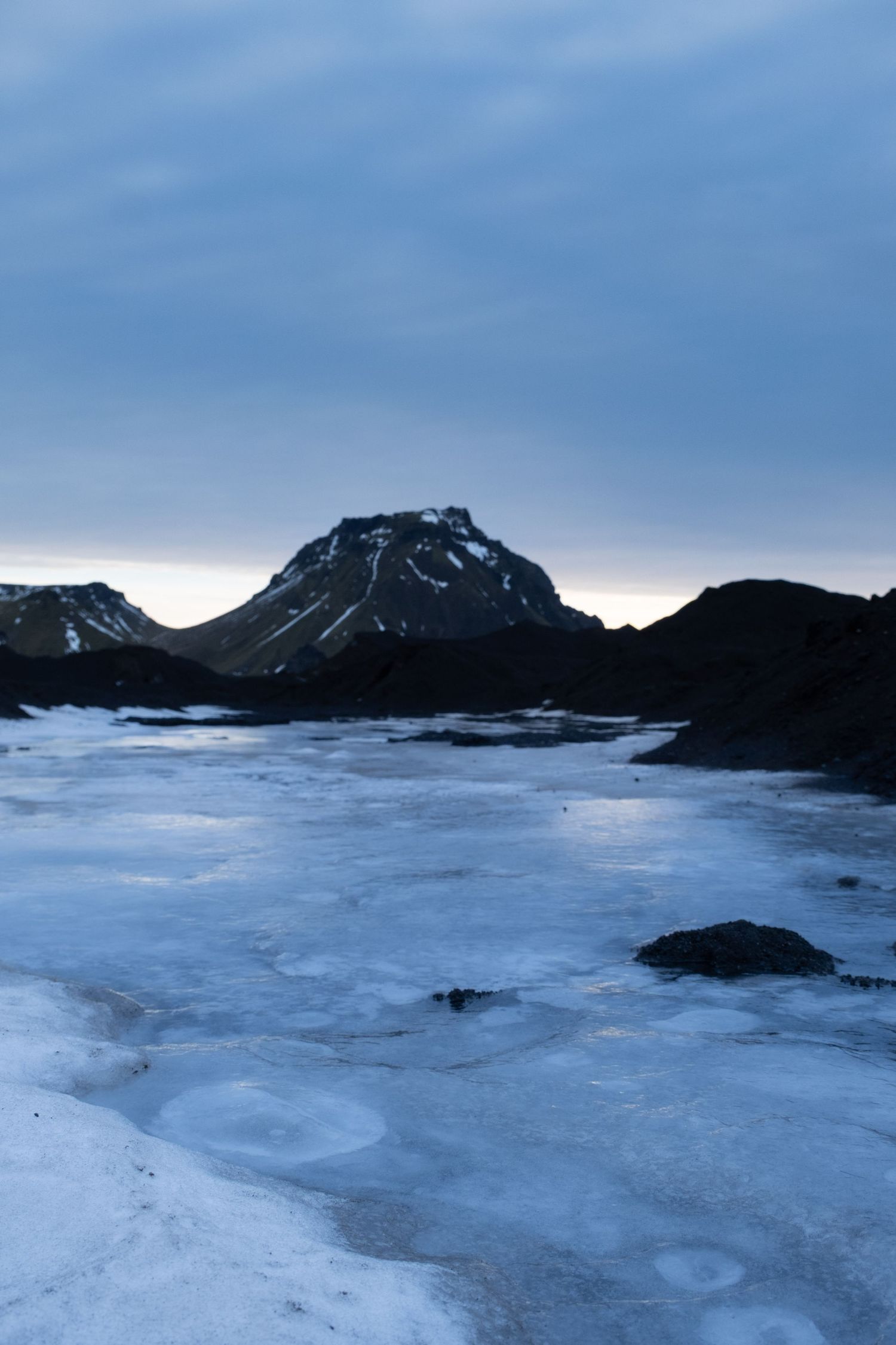 A panoramic view of snow-covered mountains and frozen lake under cloudy skies in a desolate winter landscape.