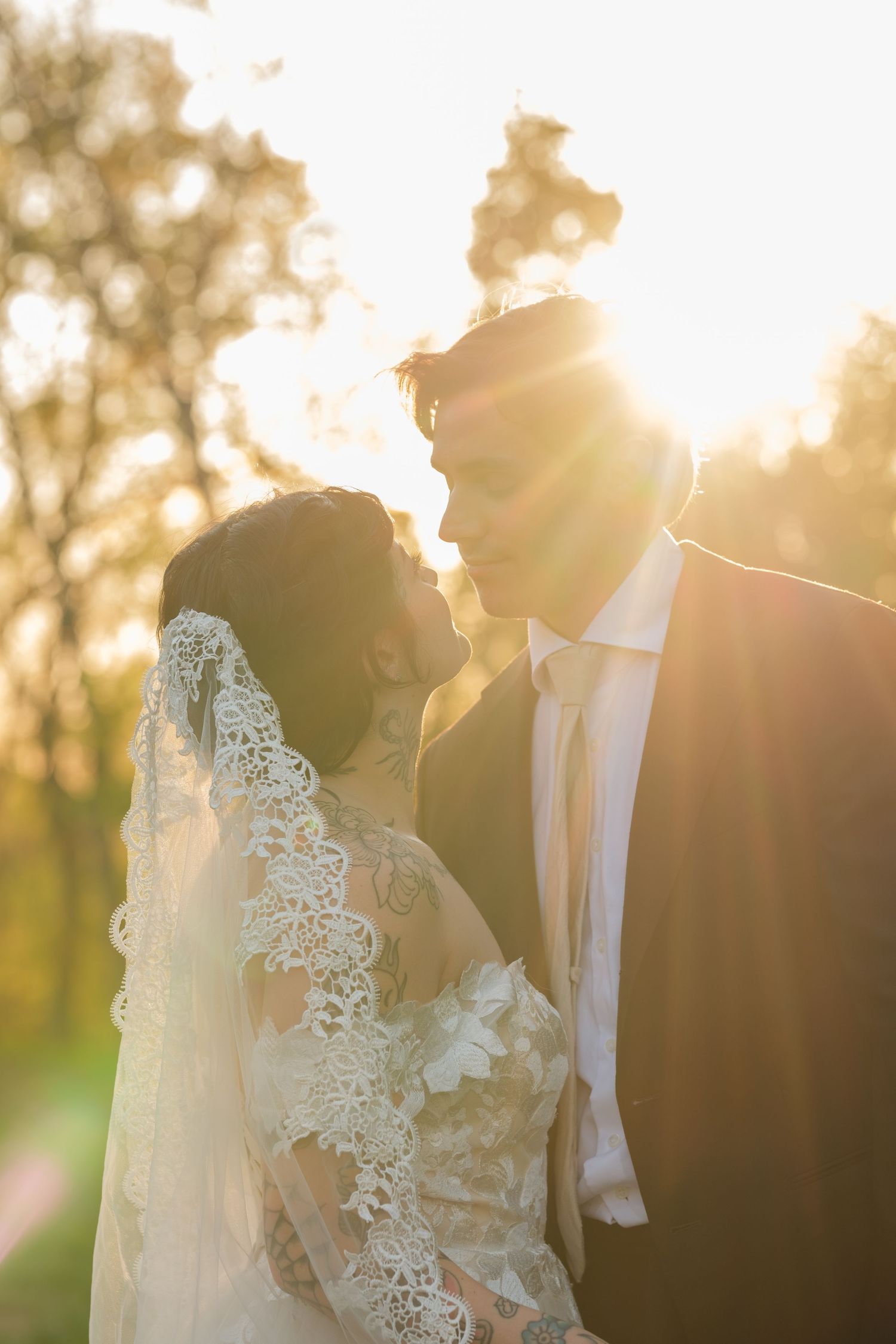 Bride and groom share a romantic moment at golden hour, with sun flare illuminating her lace veil outdoors.