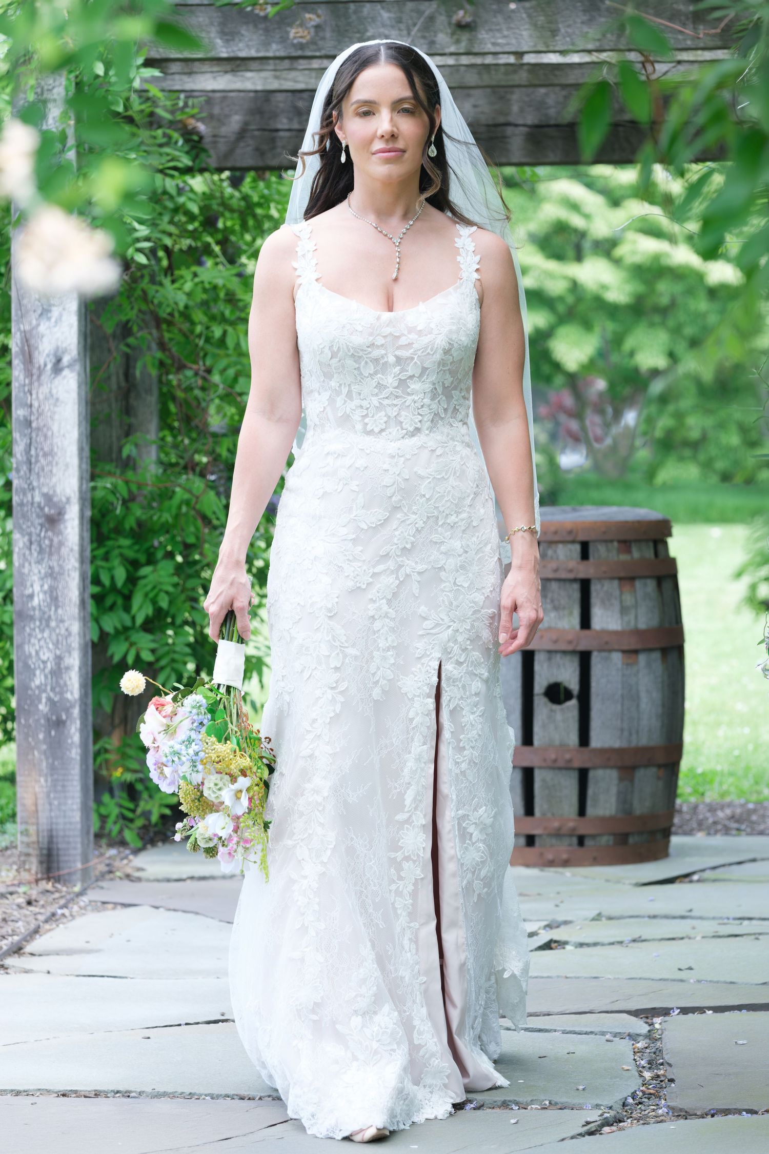 Bride in white lace gown with veil holding bouquet, walking through garden arbor with wooden barrel backdrop.