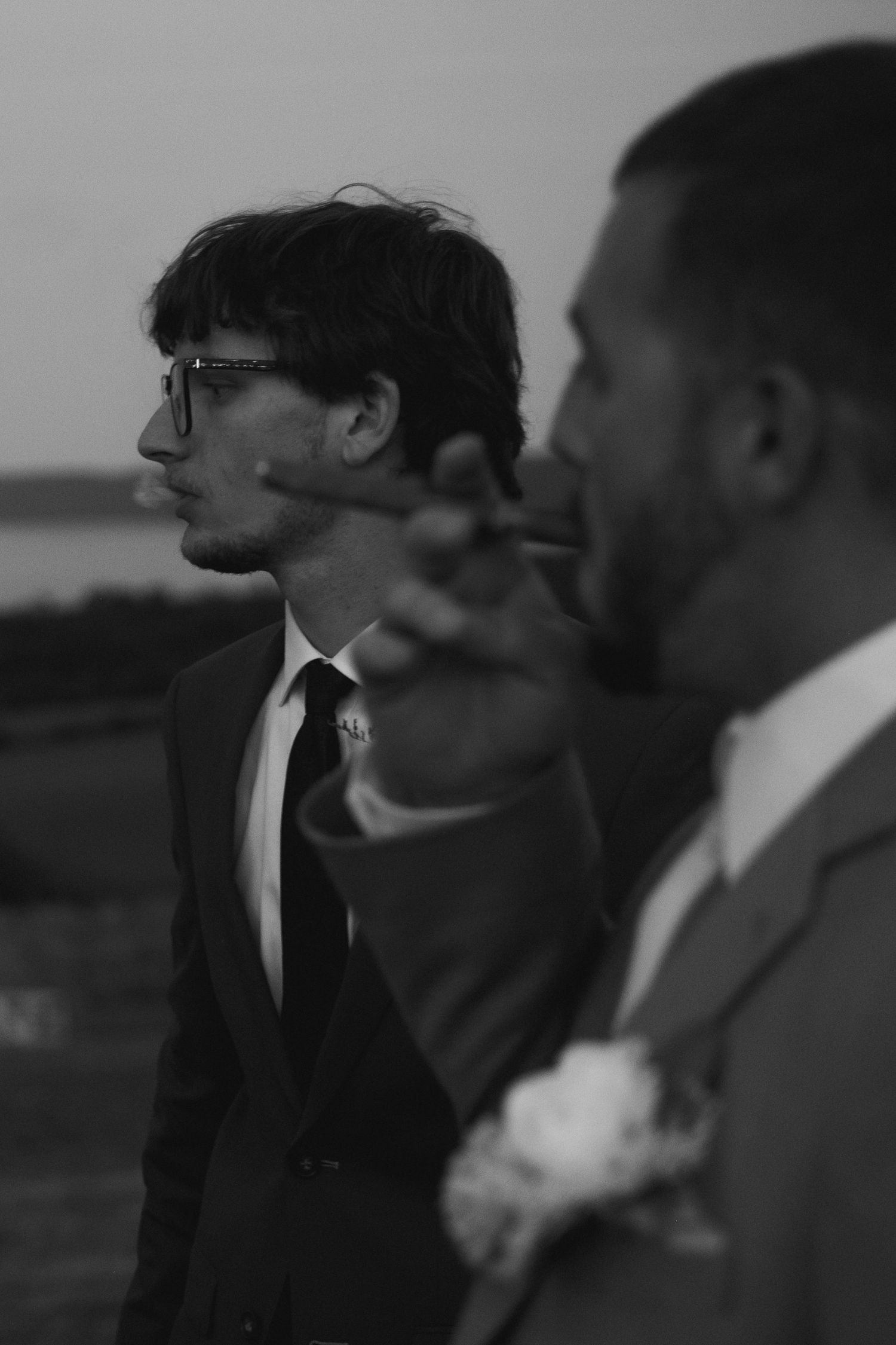 Two grooms in suits share a cigar outdoors at their wedding reception in a black and white photo.