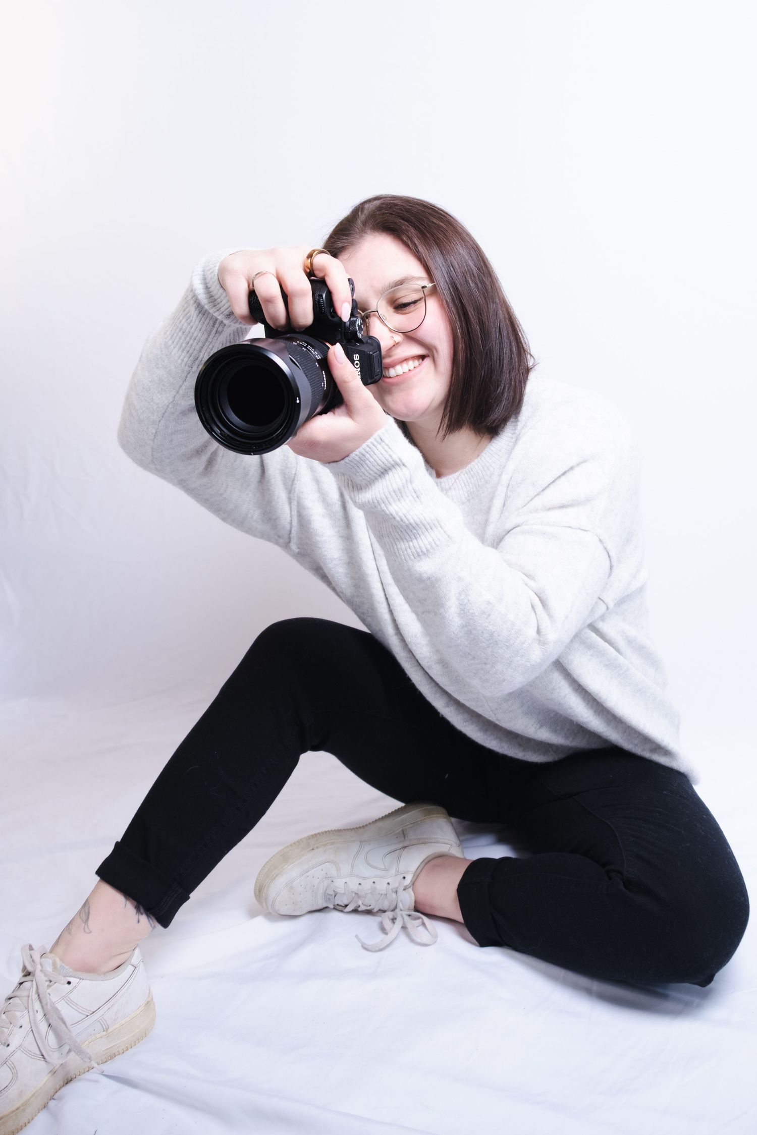 A photographer in a gray sweater and black pants lays on the floor while taking photos with a professional camera.