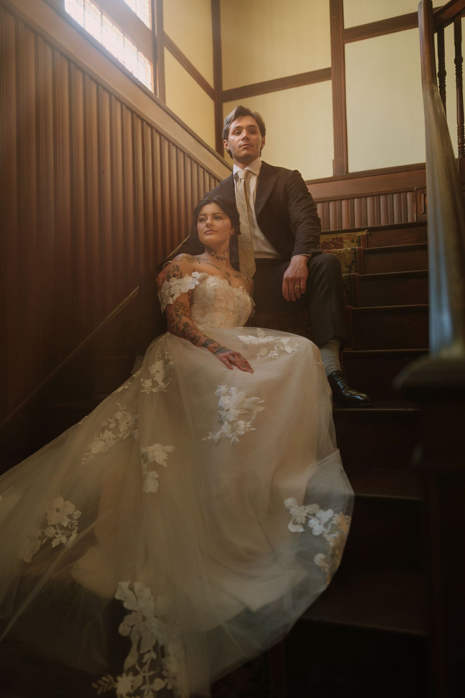 Bride in floral lace gown sits on wooden staircase beside groom in dark suit, bathed in warm window light.