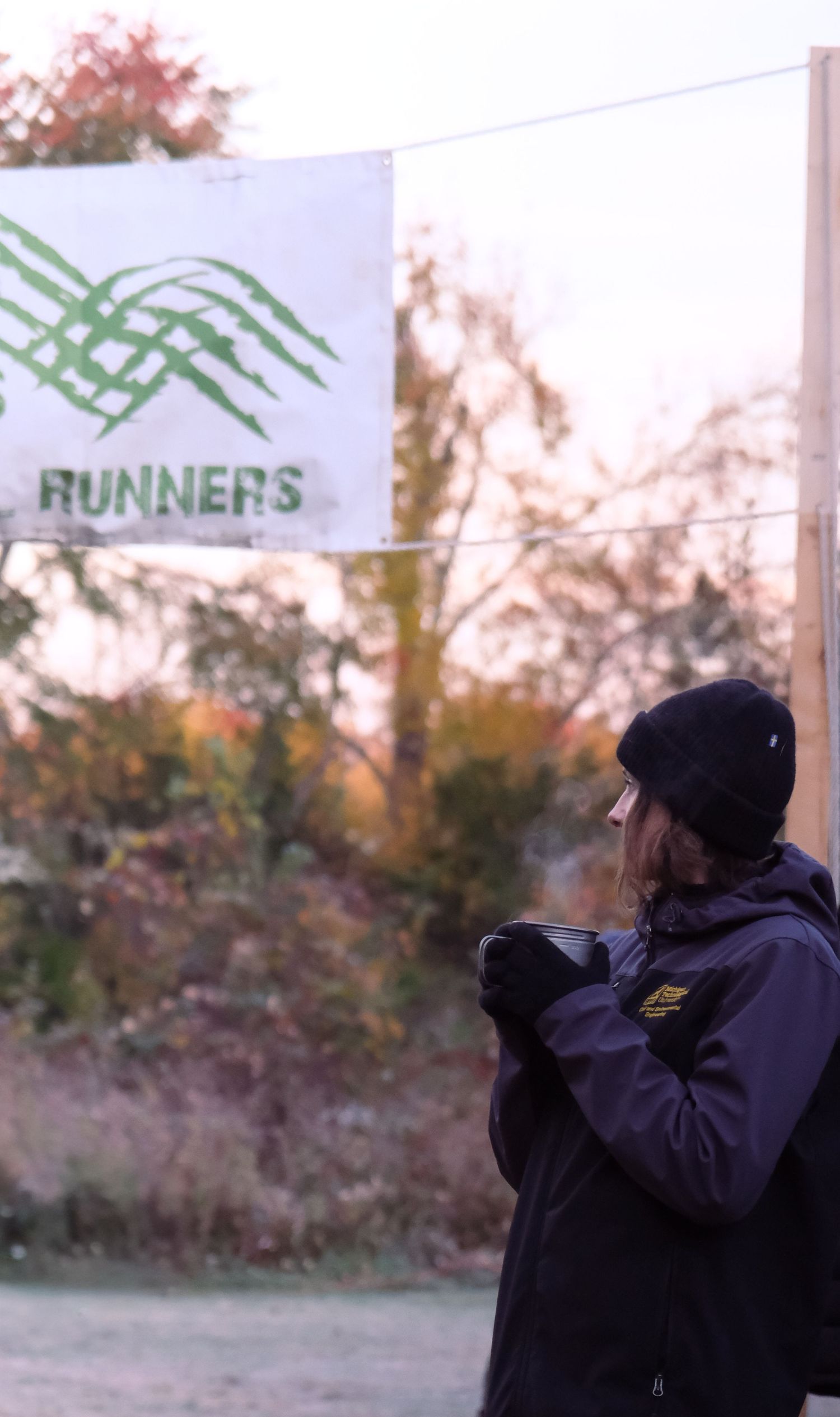 A person in a black beanie and jacket holds a hot beverage outdoors near a sign with green text reading 'Runners'.